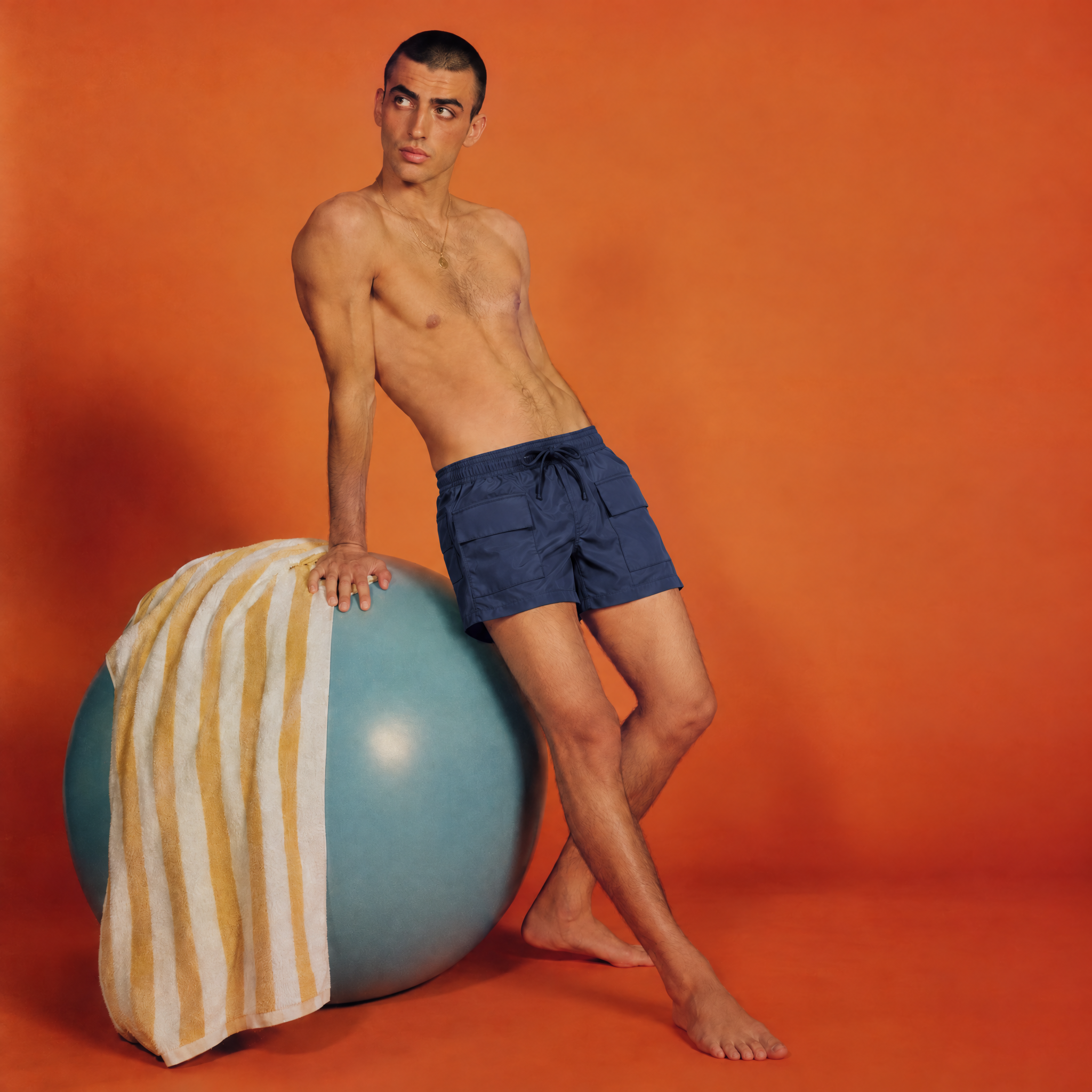 Fashion editorial featuring a model wearing Granite Island MONTECITO navy blue swim shorts against a vibrant orange studio background.