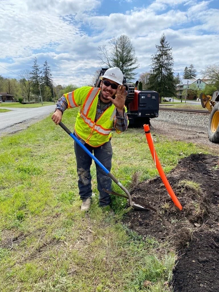 Construction worker smiling and waving, wearing a hard hat, sunglasses, and a high-visibility vest, standing next to a trench with equipment and a construction machine in the background.