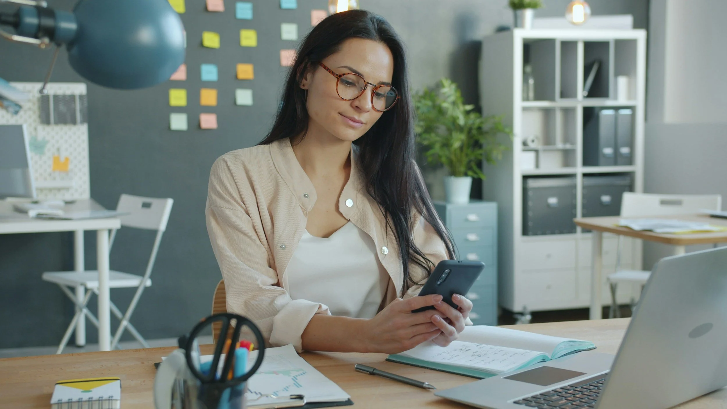A woman with long dark hair and glasses sitting at a desk in an office, looking at her smartphone, with office supplies and a laptop on her desk.