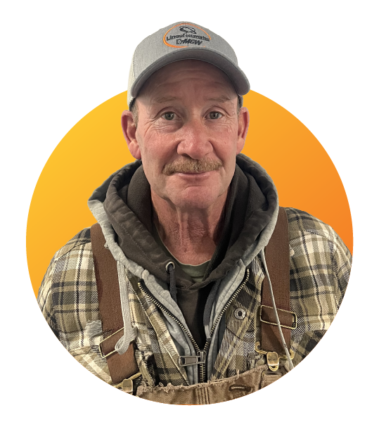 A man wearing a gray baseball cap, a plaid jacket, and layered clothes standing in front of a yellow and orange background.