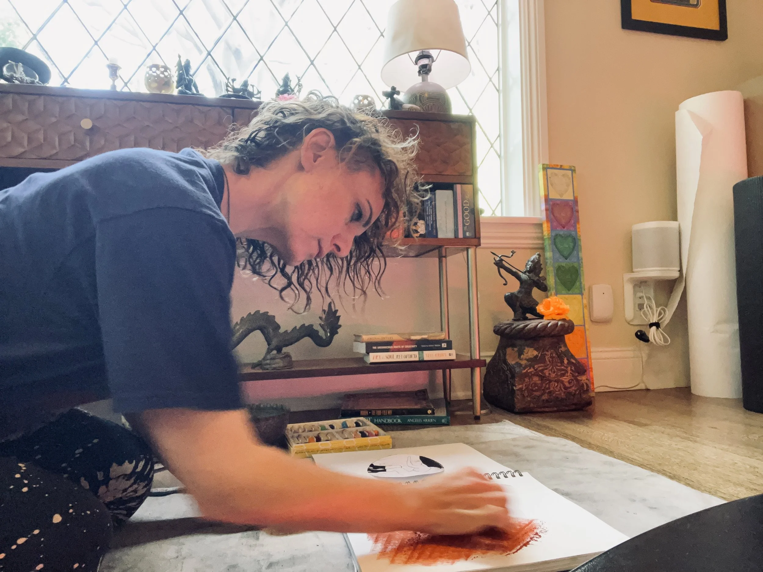 Child drawing on paper with colored pencils or markers on a table in a cozy room, with books, artwork, and decorations in the background.