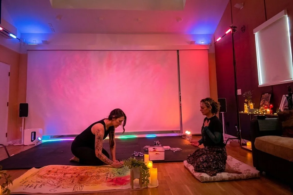 Two women sitting on the floor in a dimly lit room with colorful mood lighting, candles, and a backdrop. One woman has tattoos and is kneeling, while the other holds a small object and has curly hair. The room is decorated with plants and small lighted objects.