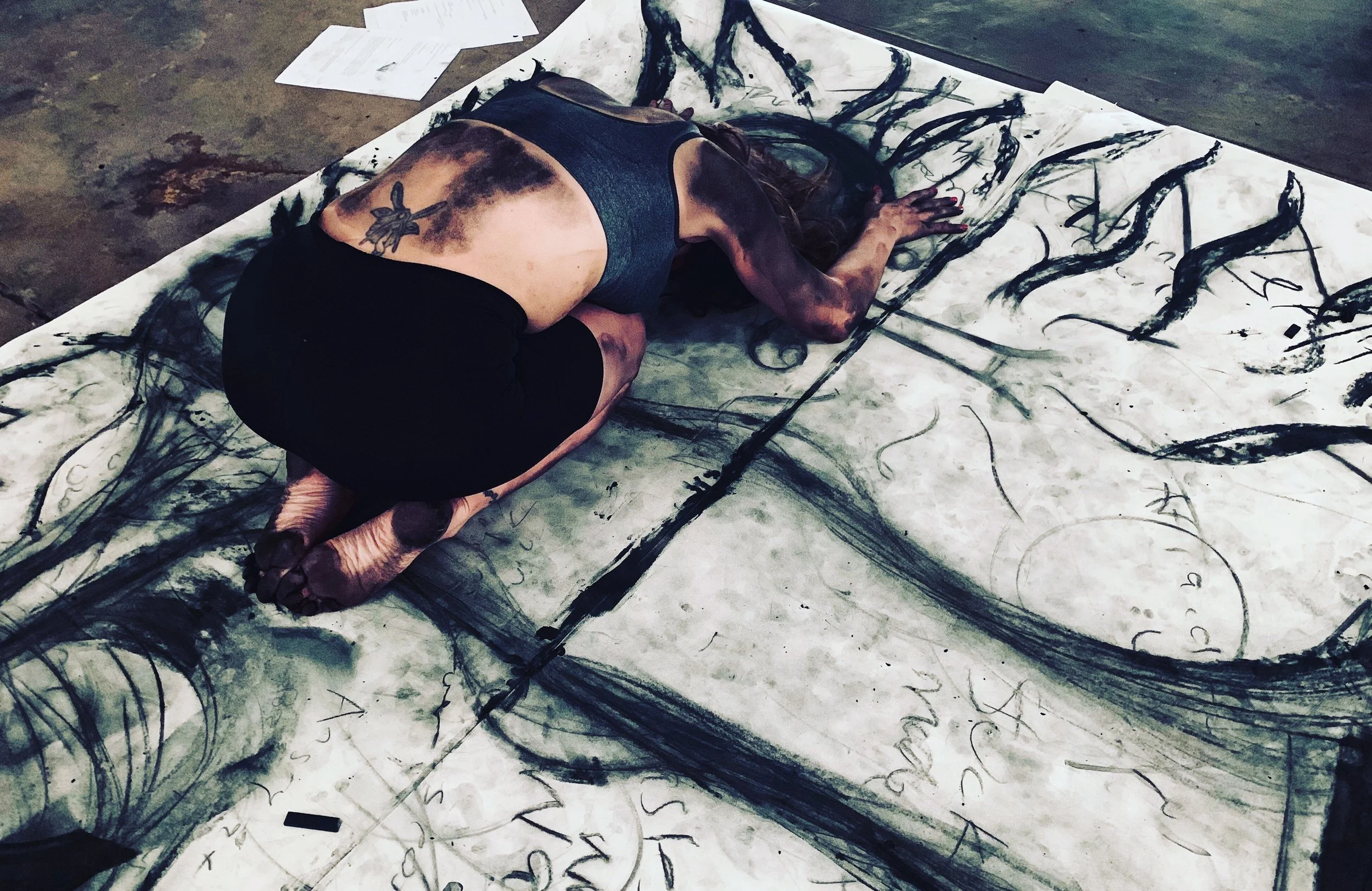 A woman with dark hair, tattoos on her arm, and wearing a sleeveless top, is kneeling on the floor and painting with black ink on large sheets of paper laid on the ground. The artwork features bold, expressive black strokes and abstract shapes.