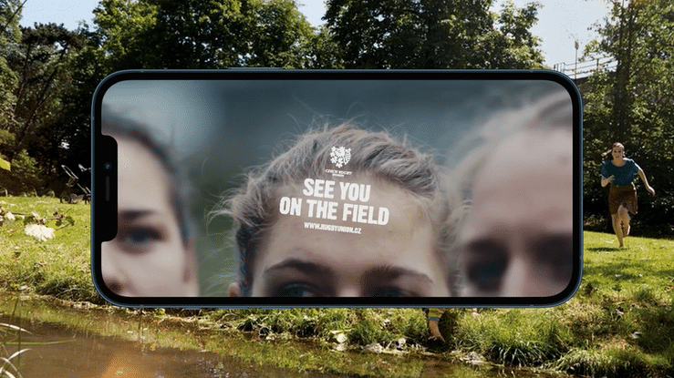 Close-up of a smartphone held above a landscape, displaying a blurred close-up of three girls' faces, with text saying 'SEE YOU ON THE FIELD' and a website URL.