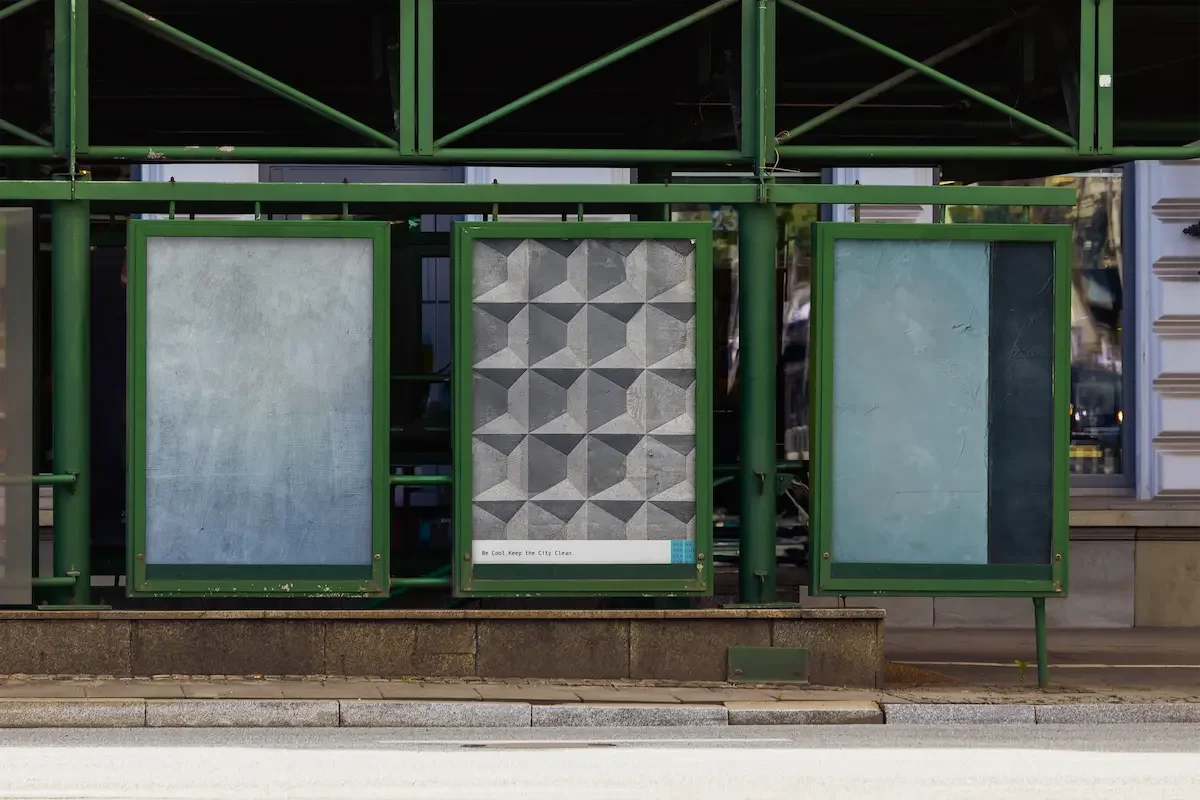 Three empty advertisement display boards mounted on a green metal structure on a city sidewalk, with a building facade in the background.