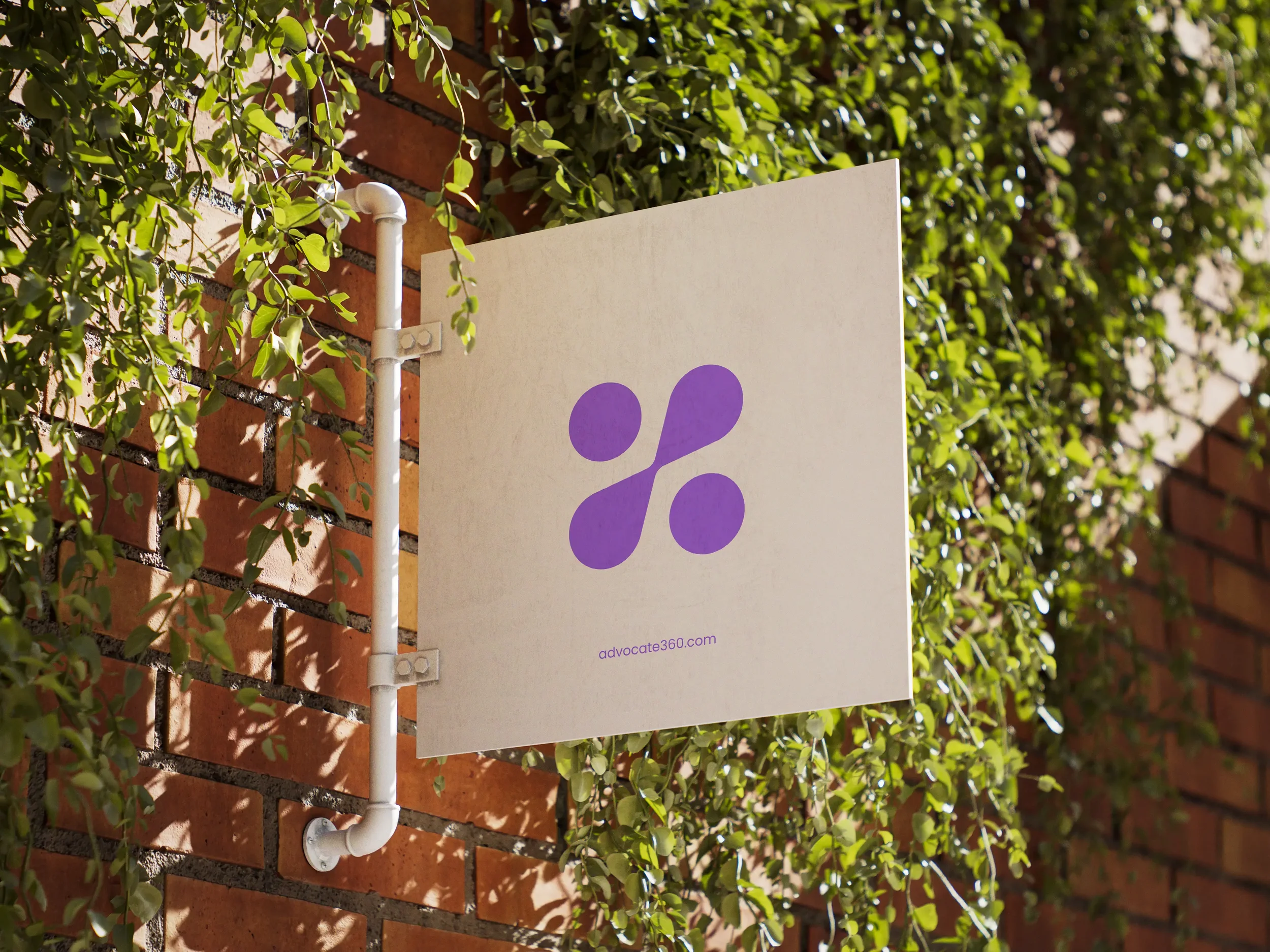 A square sign with a purple abstract logo and the website advcate360.com, mounted on a brick wall amidst green foliage.
