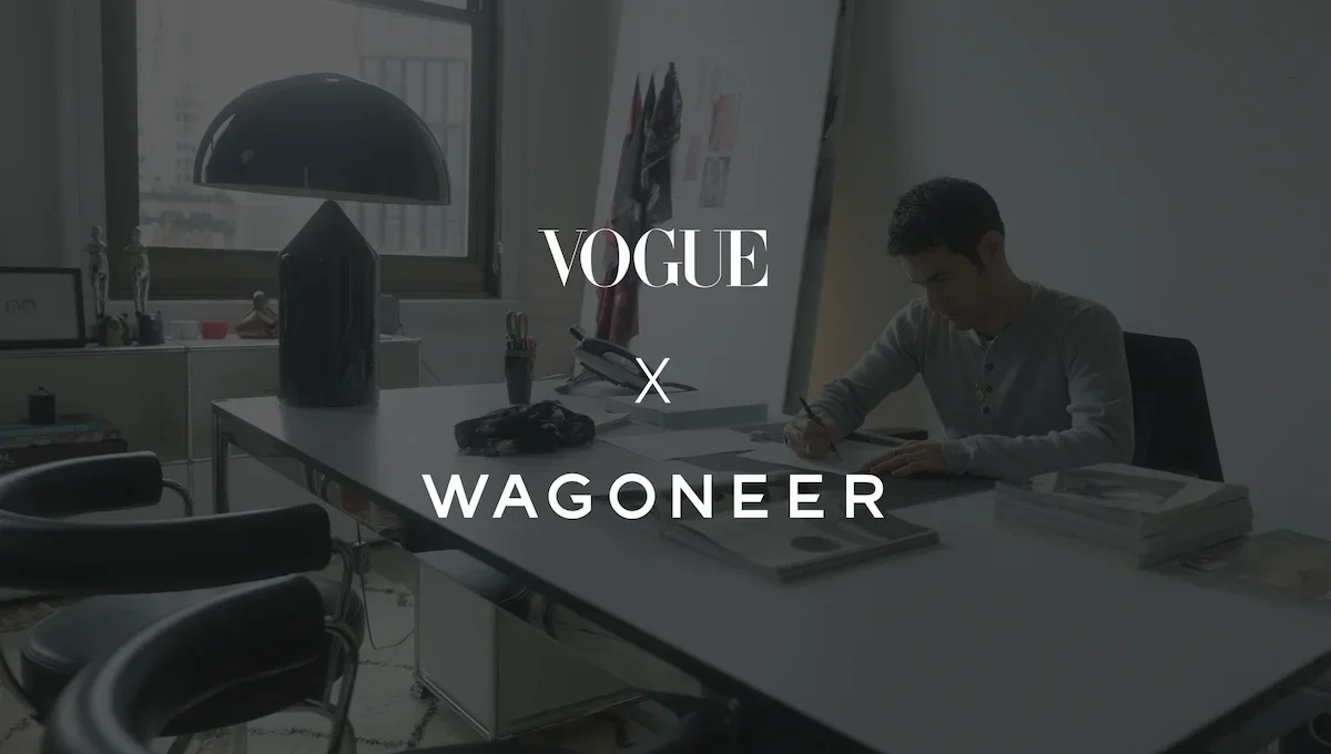 An office with a desk, a man writing, a large black desk lamp, a window, and various items on the desk and shelves, with overlay text reading 'VOGUE x WAGONER'.