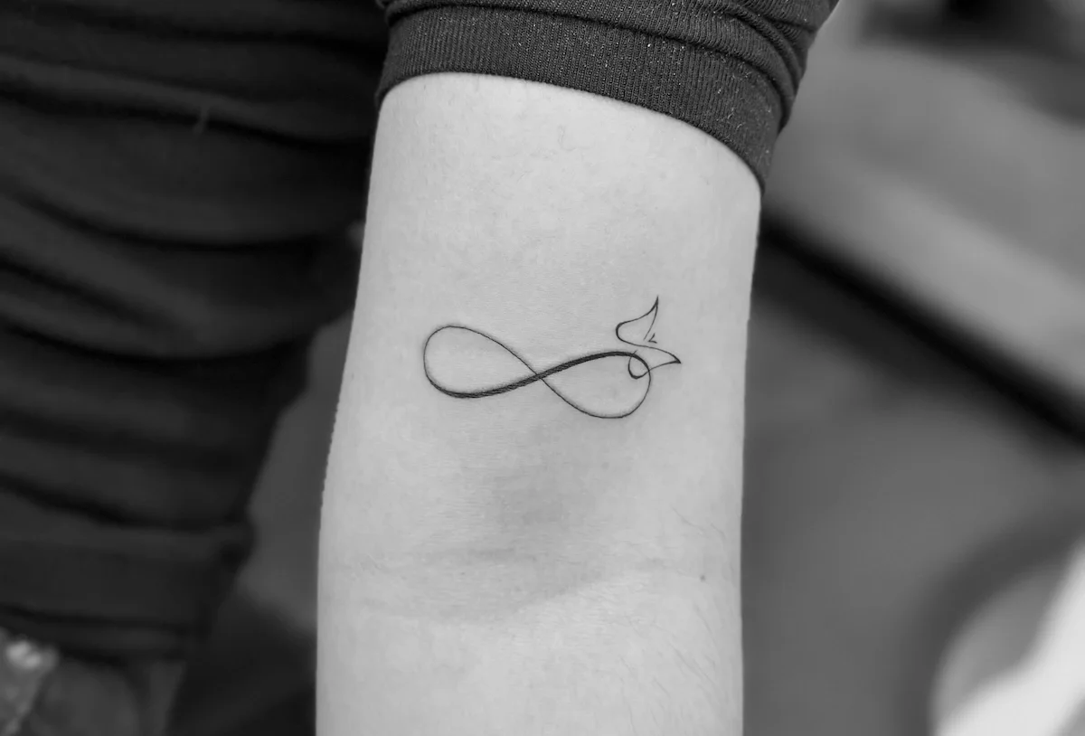 A person's arm with a small, minimalist tattoo of an infinity symbol with a small sailboat integrated into the design.