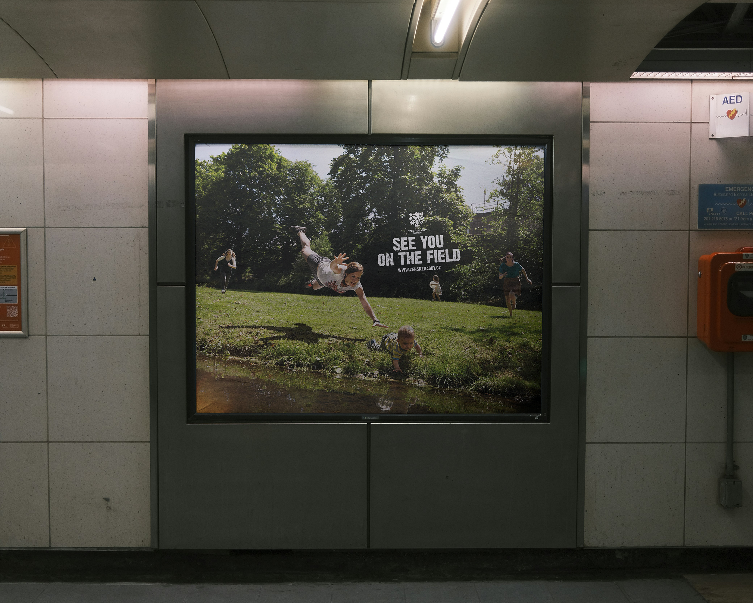 An advertisement in a subway station showing children playing outside in a grassy park with trees. The text reads "See you on the field" and includes a website address.