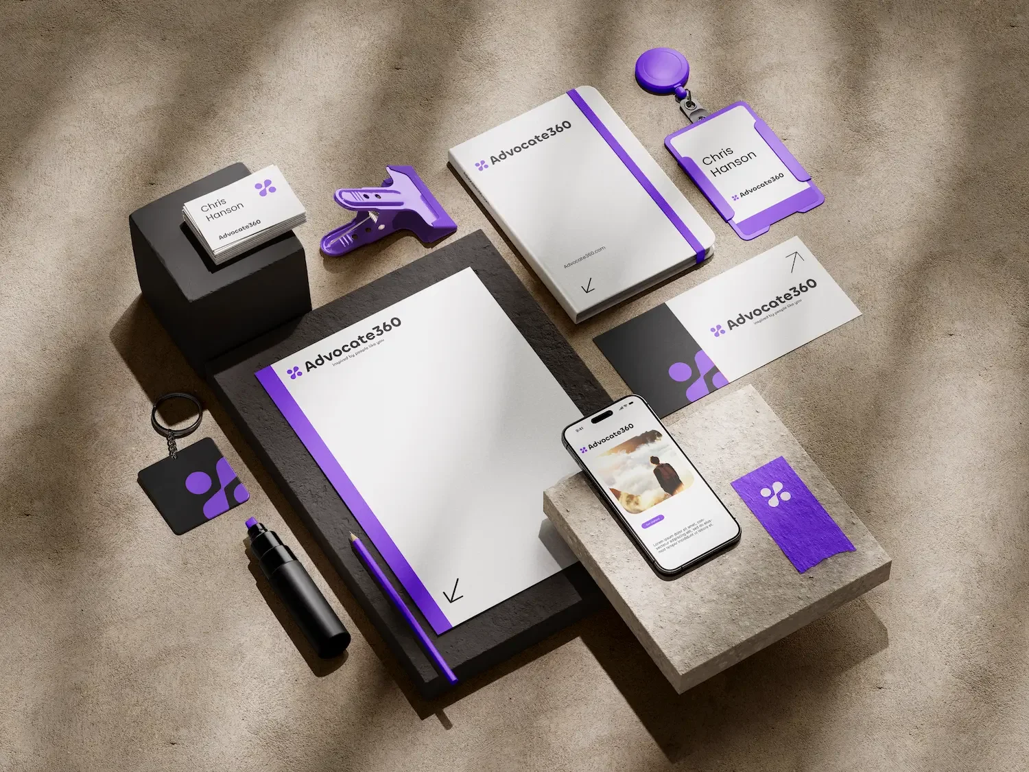 Various branded office supplies and tech devices on a beige surface, including business cards, notebooks, a smartphone, a badge, and a marker, all with a purple and white color theme for Advocate360.