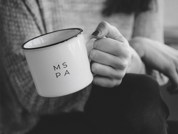 A person holding a white ceramic mug with the letters 'MS PA' printed on it, in black, in black and white photo.