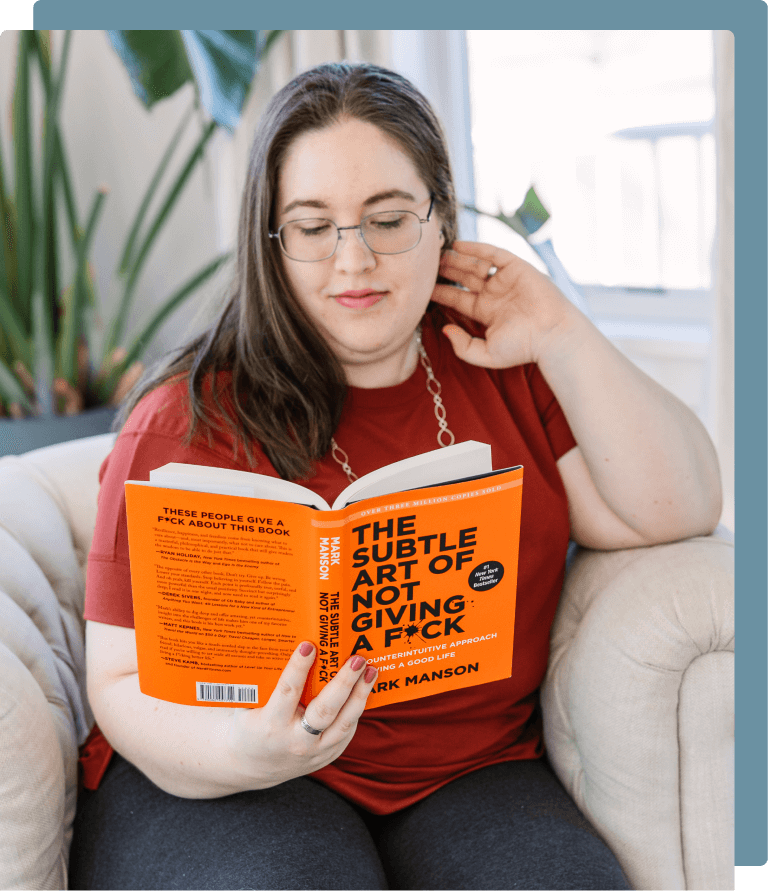 Sarah sitting on a couch in a bright living room, reading ‘The Subtle Art of Not Giving a F*ck’ by Mark Manson