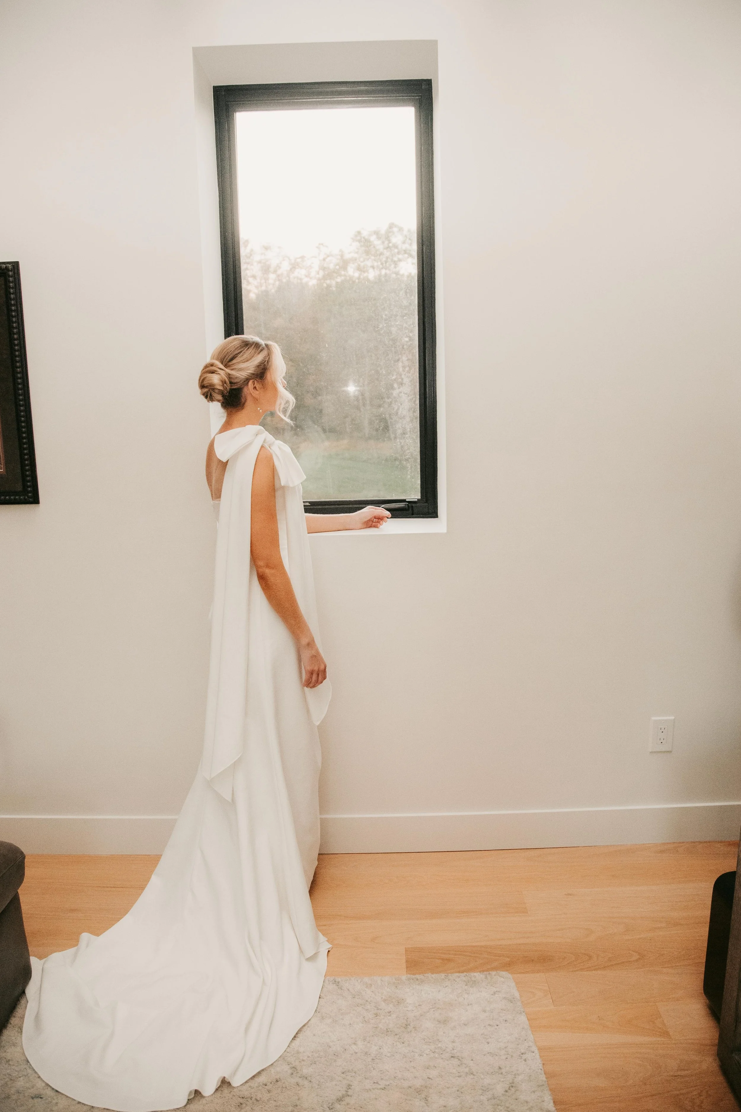 bride in simple elegant gown with draping and bow over one shoulder looks out the window
