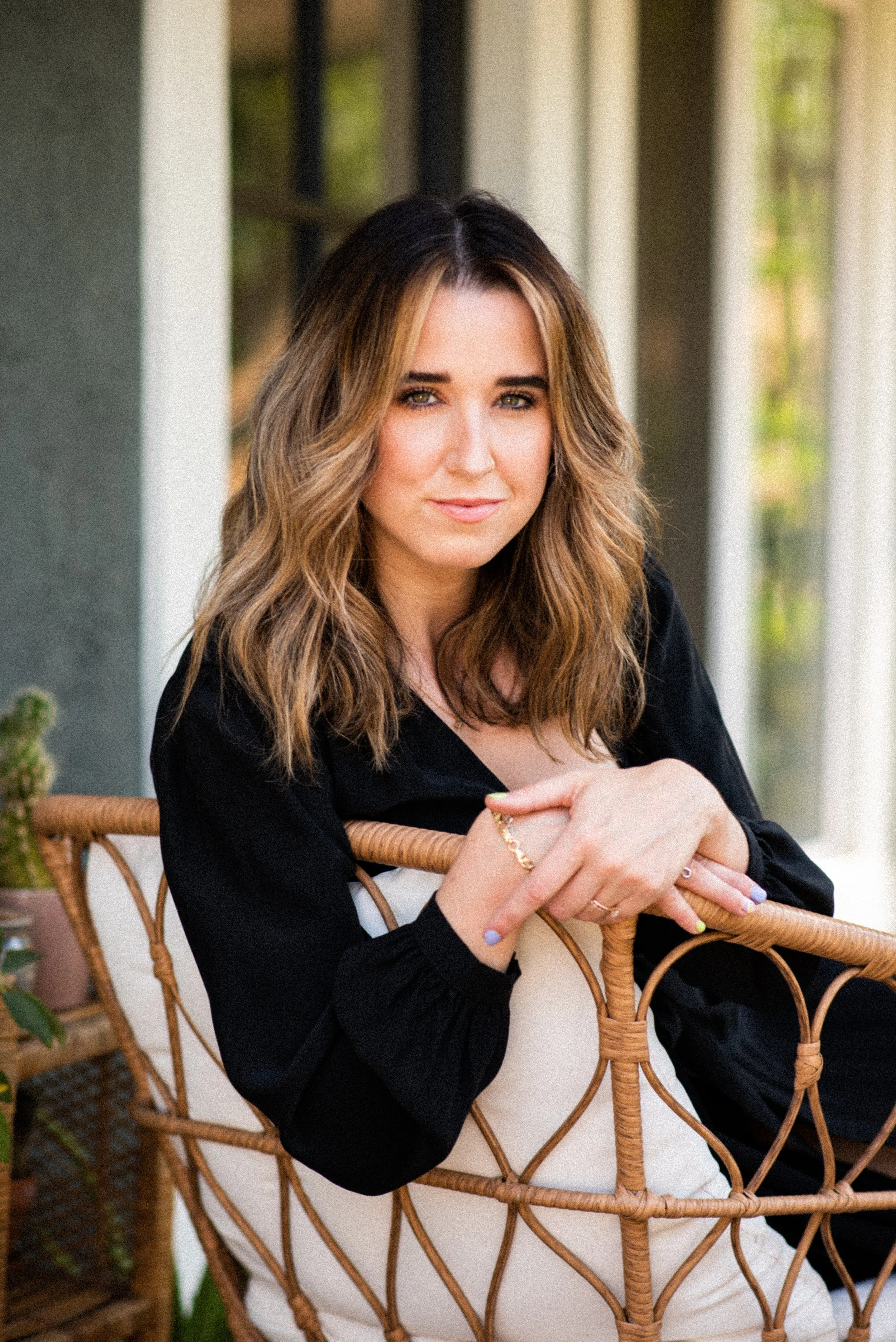 A woman with shoulder-length wavy hair, wearing a black top, sitting on a wicker chair indoors or on a covered porch, with a cautious expression. Behind her is a white wall and a window with green trees visible through it.