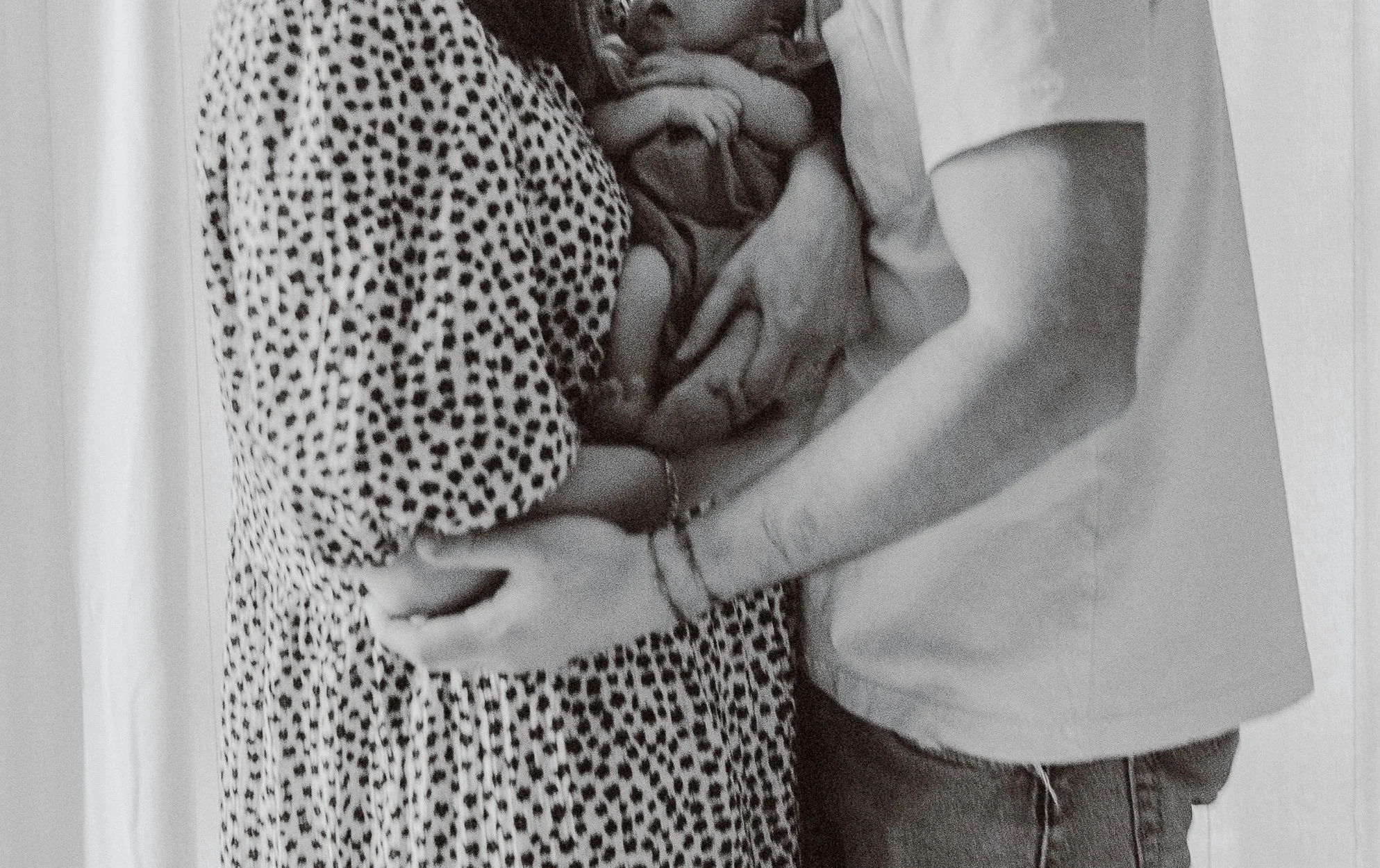 A person wearing a polka dot shirt holding a sleeping child dressed in a hoodie and shorts, both resting on a surface.