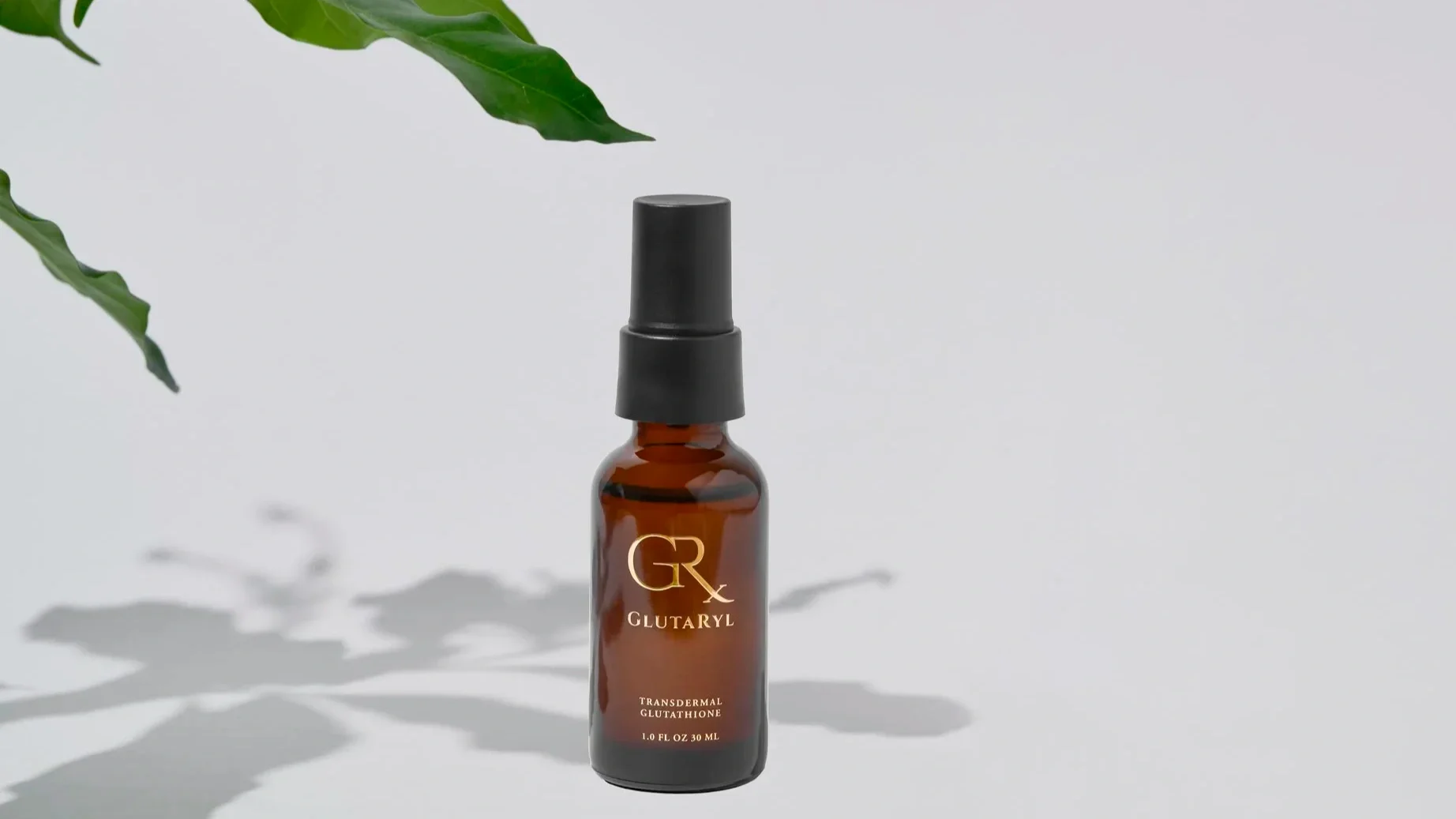 A small amber glass bottle with a black spray cap, labeled 'GR GLUTARYL, Transdermal Glutathione, 1.0 FL OZ 30 ML,' is placed on a light-colored surface with shadows, and green leaves are partially visible in the top left corner.