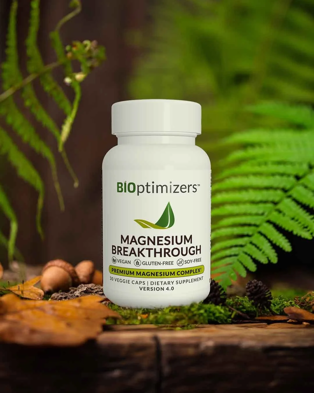 A white bottle of BIOptimizers Magnesium Breakthrough dietary supplement in front of green fern leaves and small mushrooms on a log.