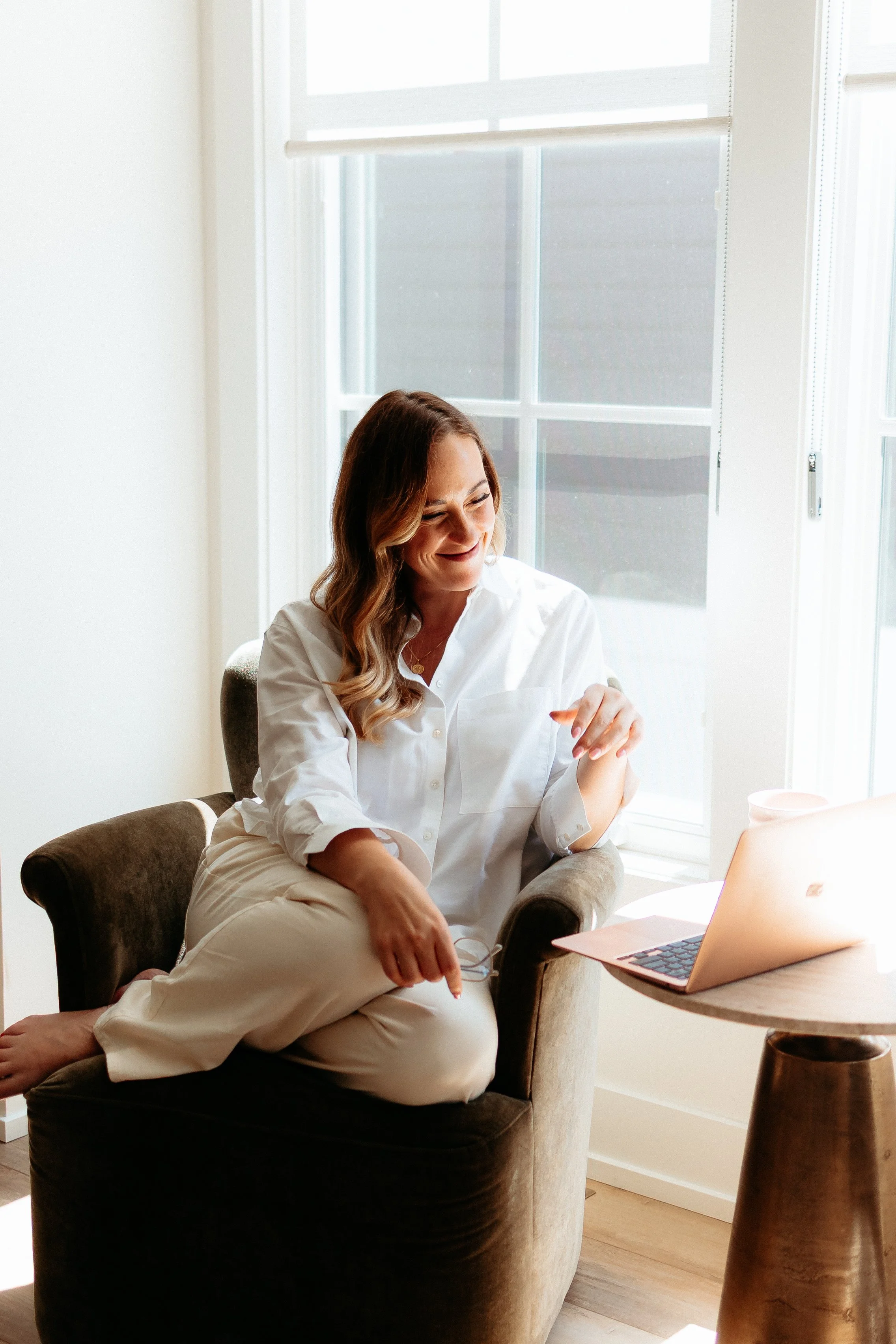 A woman with long wavy hair, wearing a white blouse and beige pants, sitting comfortably in a brown armchair near a window with white blinds, smiling and looking down at something in her hand. There is a laptop on the small round wooden table next to her.