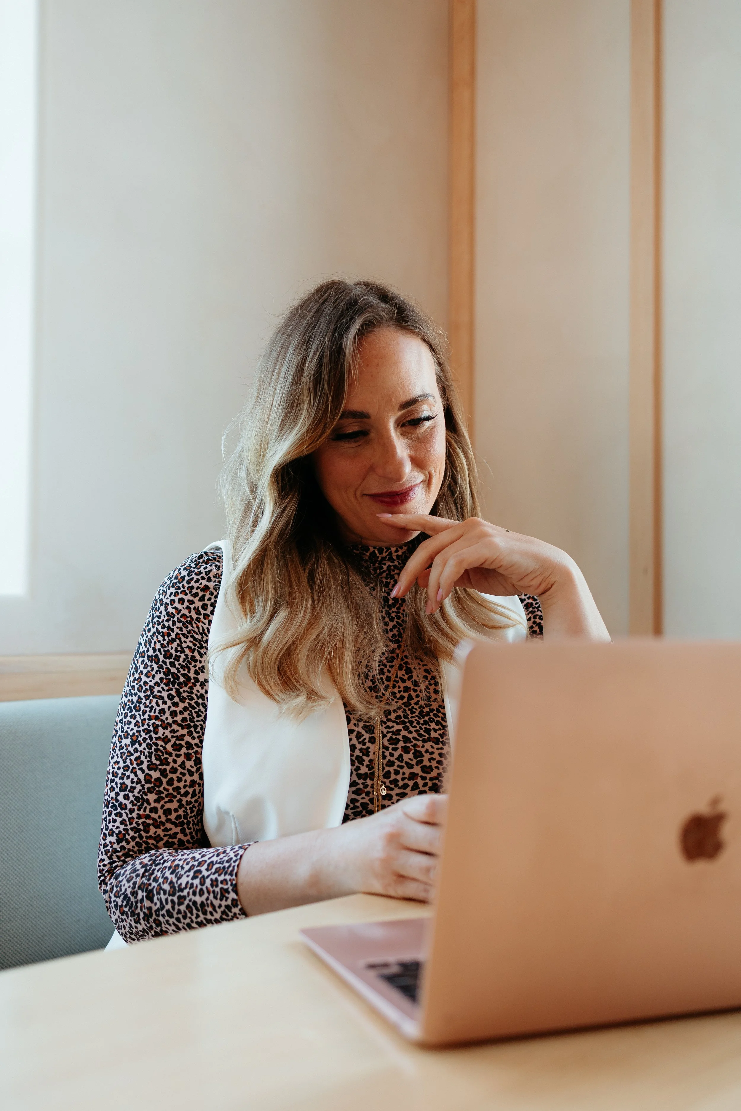 A woman with long wavy hair, wearing a leopard print top and white vest, sitting at a table and looking at a laptop.
