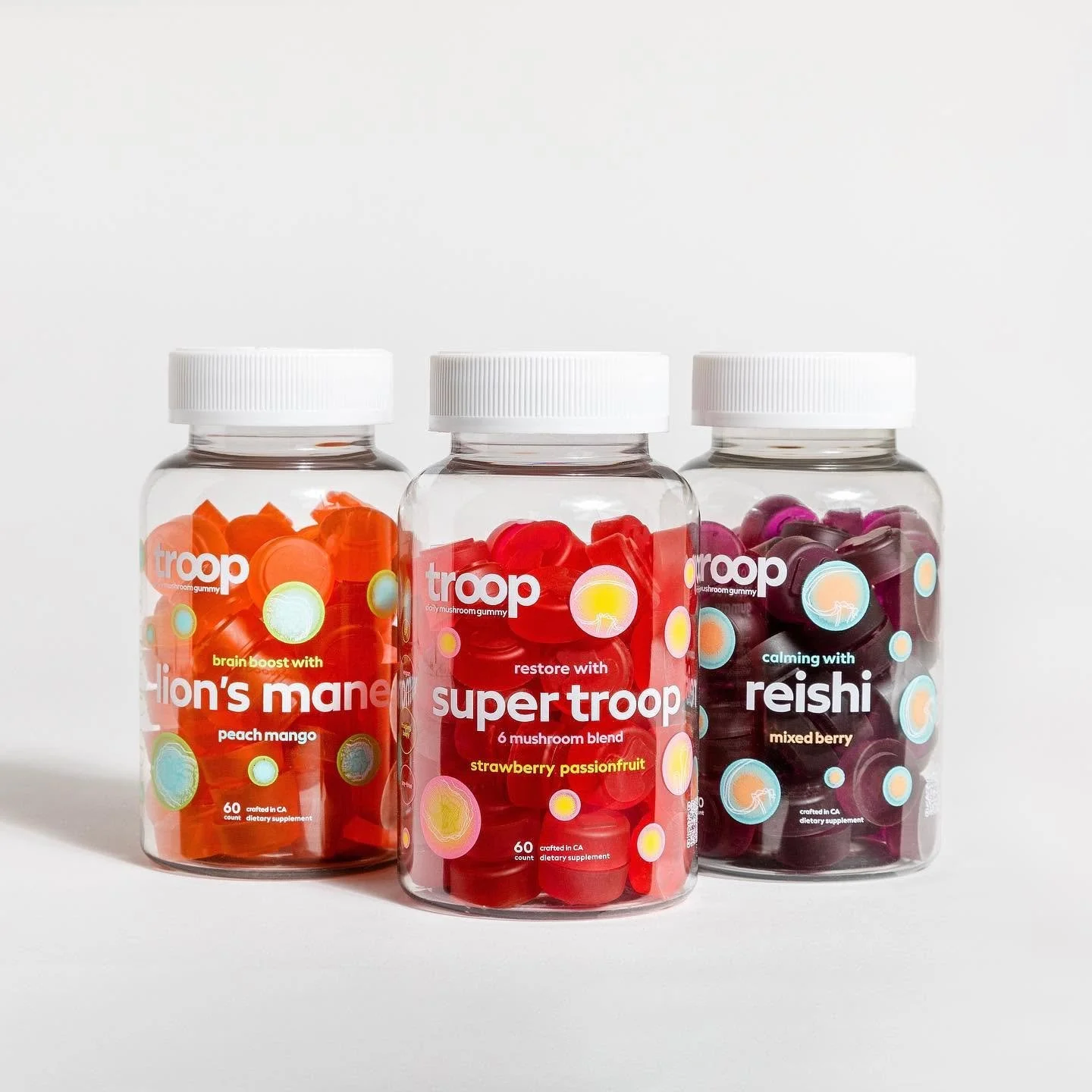 Three bottles of mushroom gummies labeled 'troop'. The left one is peach mango flavor, the middle one is strawberry passionfruit flavor, and the right one is mixed berry flavor, placed on a white background.
