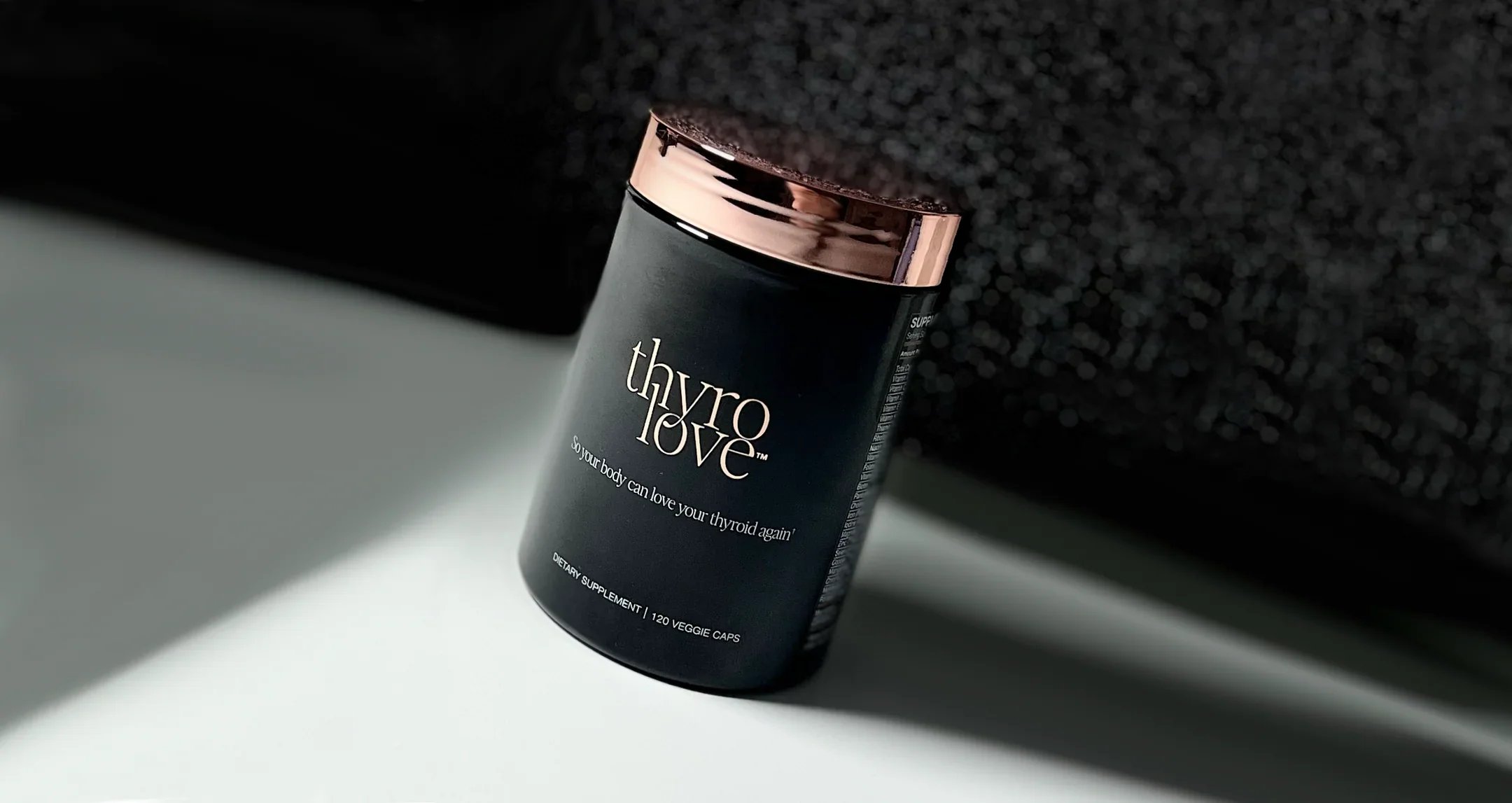 Black bottle labeled 'thyro love' with rose gold cap, positioned diagonally on a white and black textured background.