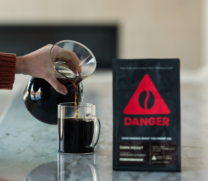 A person is pouring hot coffee from a pot into a glass mug on a kitchen countertop. There is a warning sign in the background indicating danger, likely related to the hot liquid.