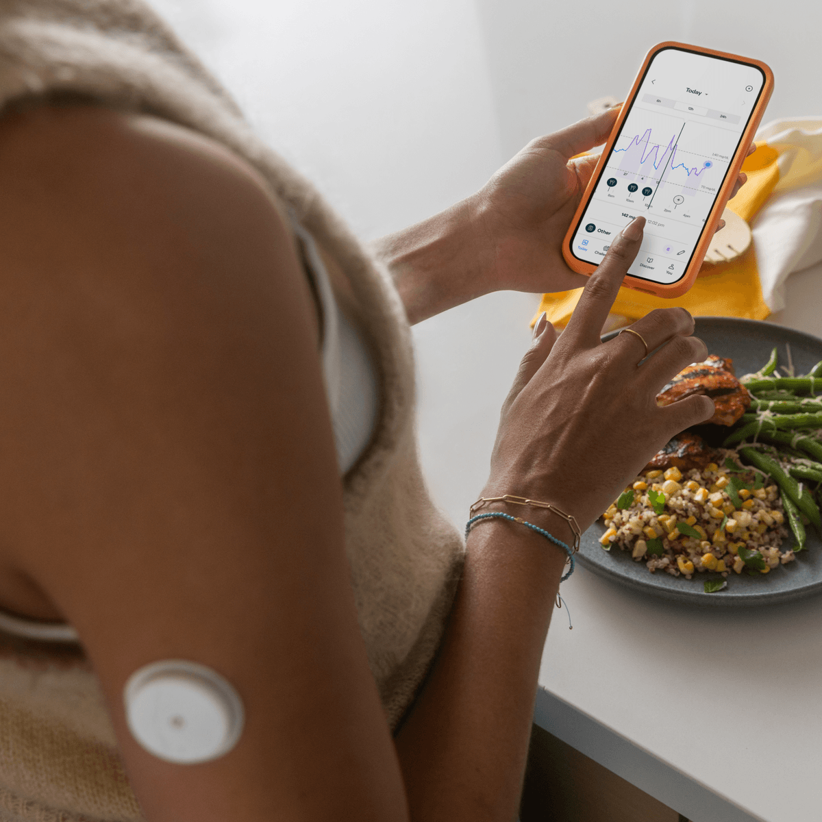A person holding a smartphone with a health tracking app open, showing a graph and data, sitting at a table with a plate of food including green beans and cooked grains.