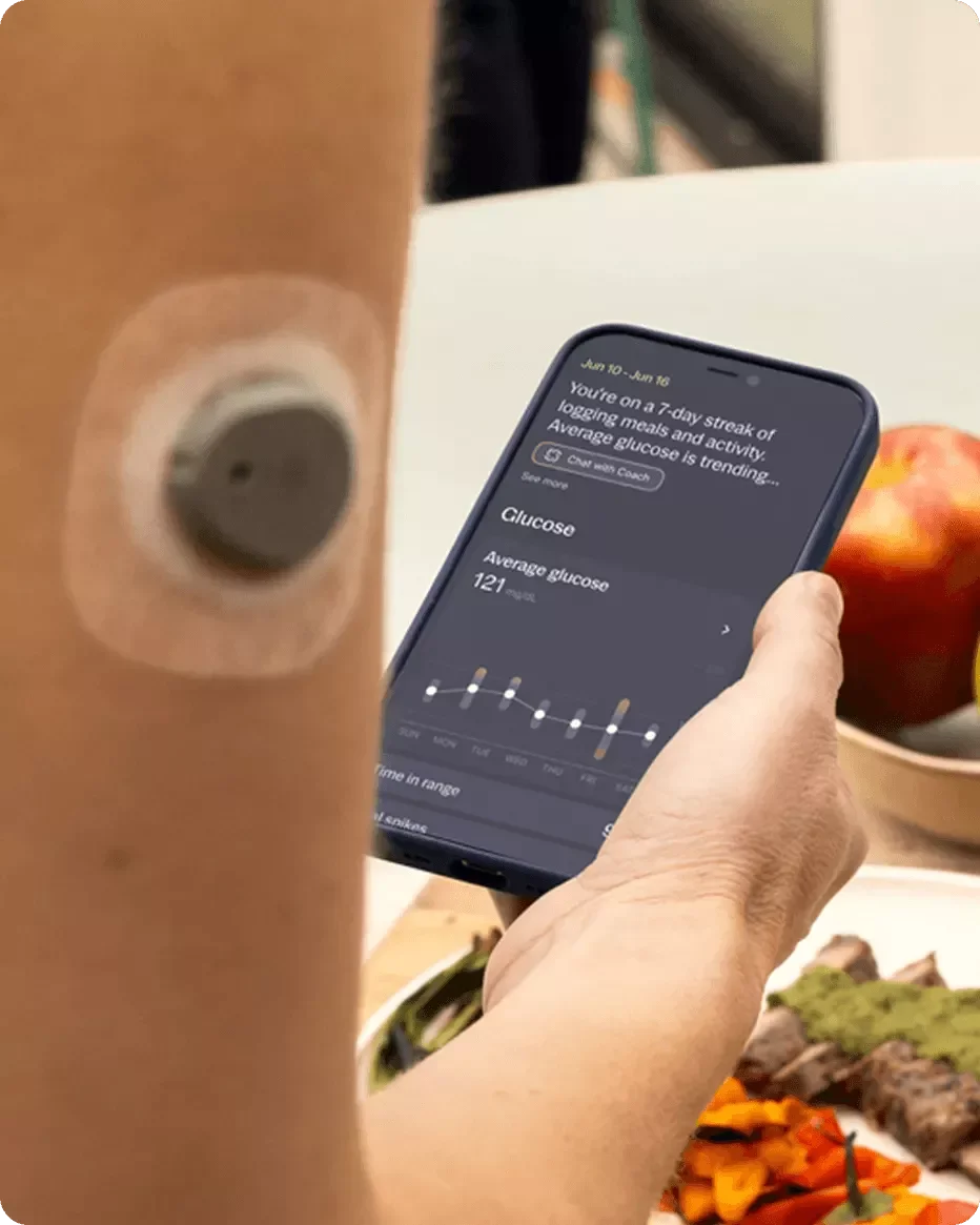 Close-up of a person's arm with a circular fitness tracker worn on their wrist, holding a smartphone displaying a health tracking app with data on glucose levels and activity. In the background, there are plates of food, including a bowl of apples and a meal with vegetables and meat.