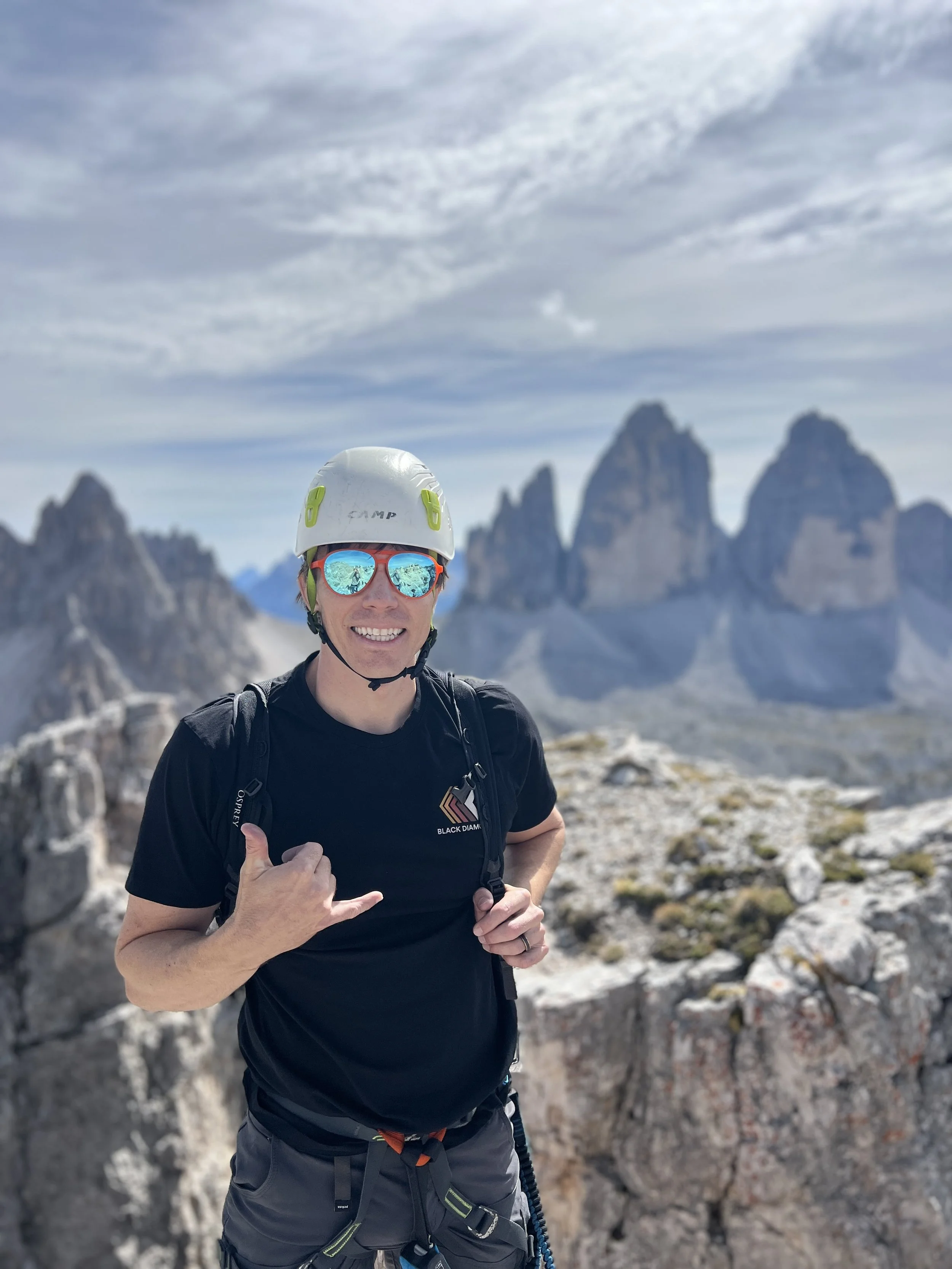 A man wearing a helmet, sunglasses, and a backpack, smiling and making a shaka sign, standing on rocky terrain with mountain peaks in the background.