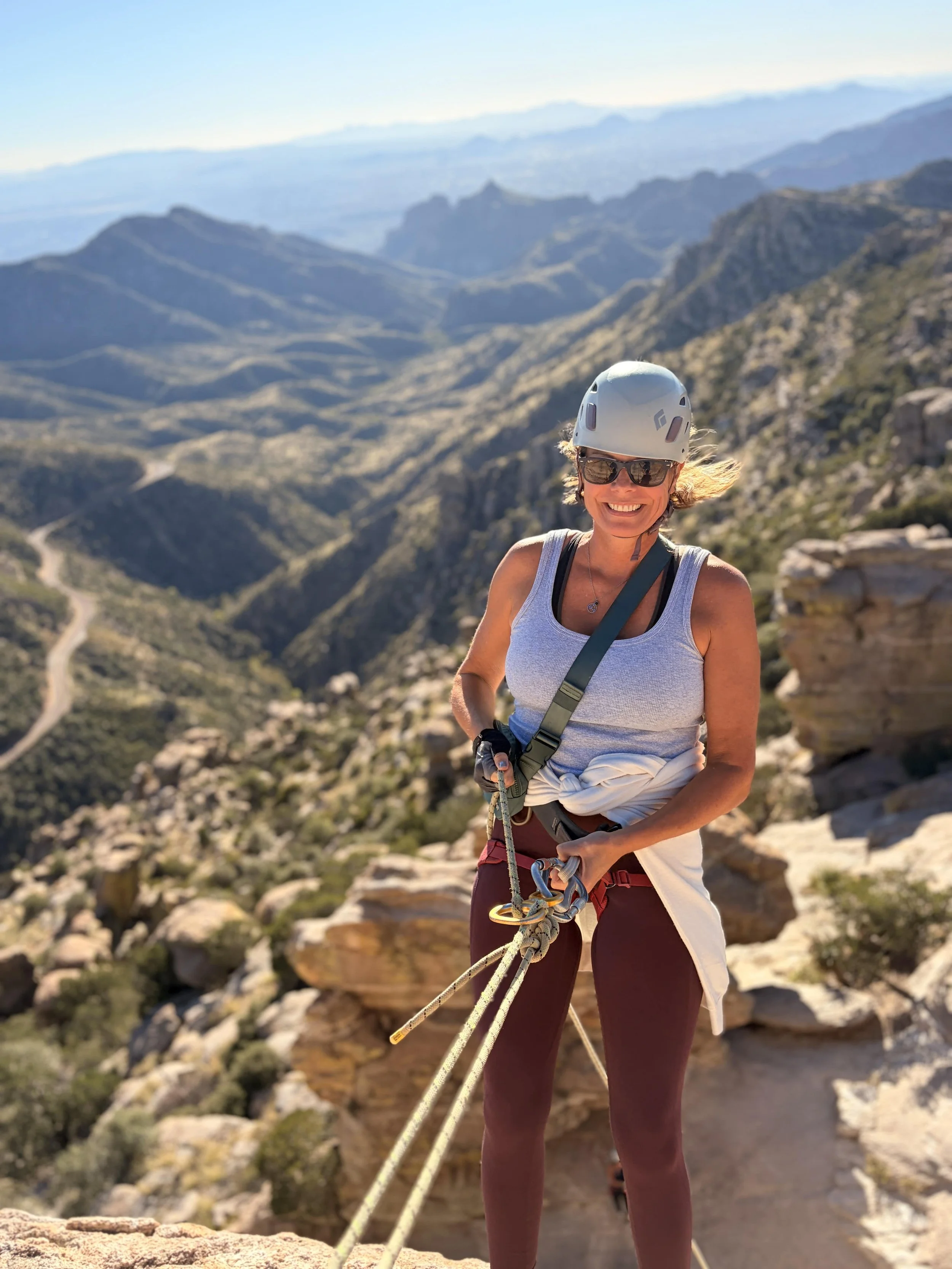 A smiling woman wearing a helmet and sunglasses standing on a rocky mountain trail, holding a climbing rope with a scenic mountain landscape in the background.
