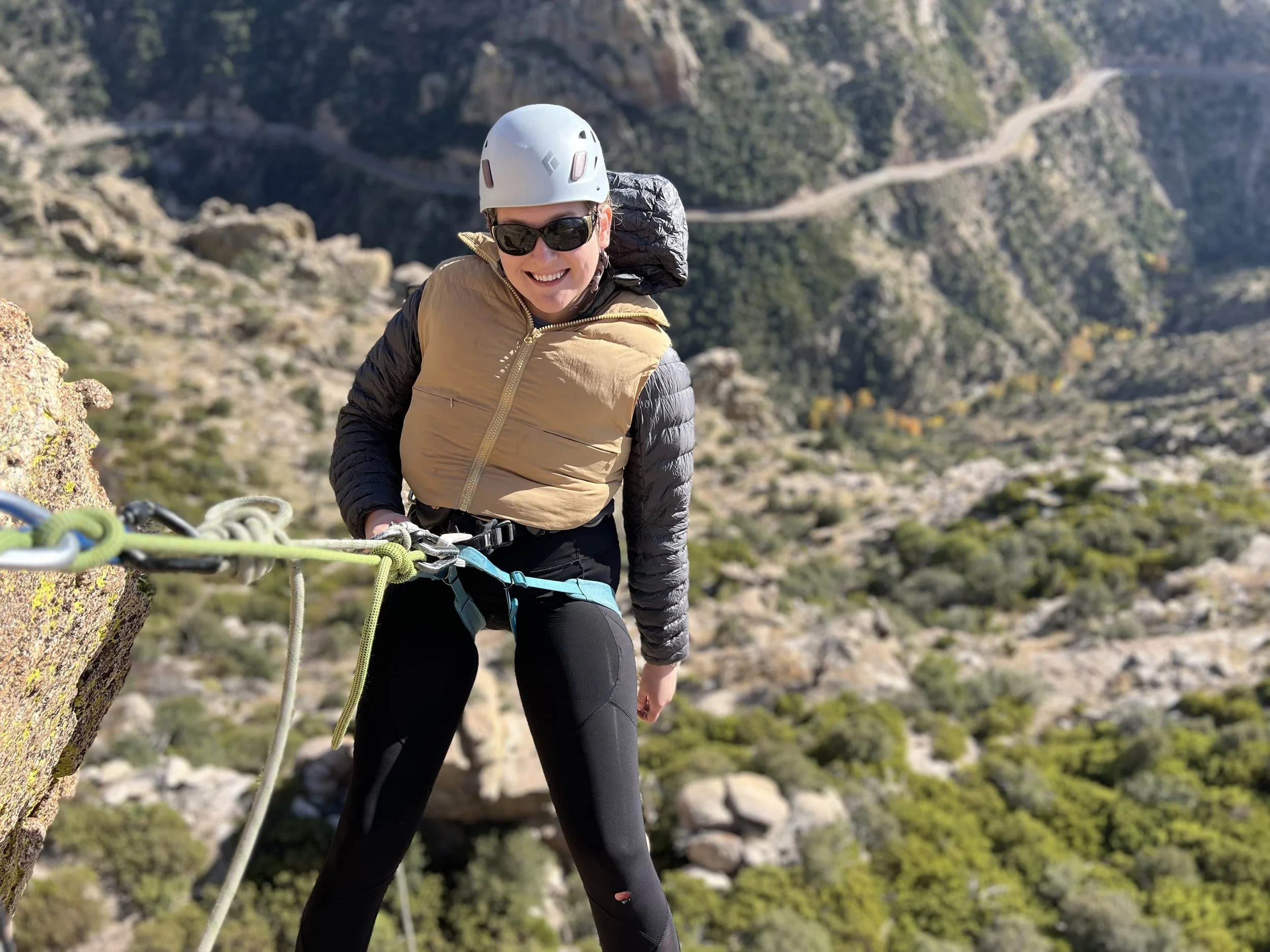 A woman in climbing gear, including a helmet black sunglasses, a beige puffy vest, and black pants, stands on a rocky mountain ledge with a scenic landscape of mountains and trees in the background.