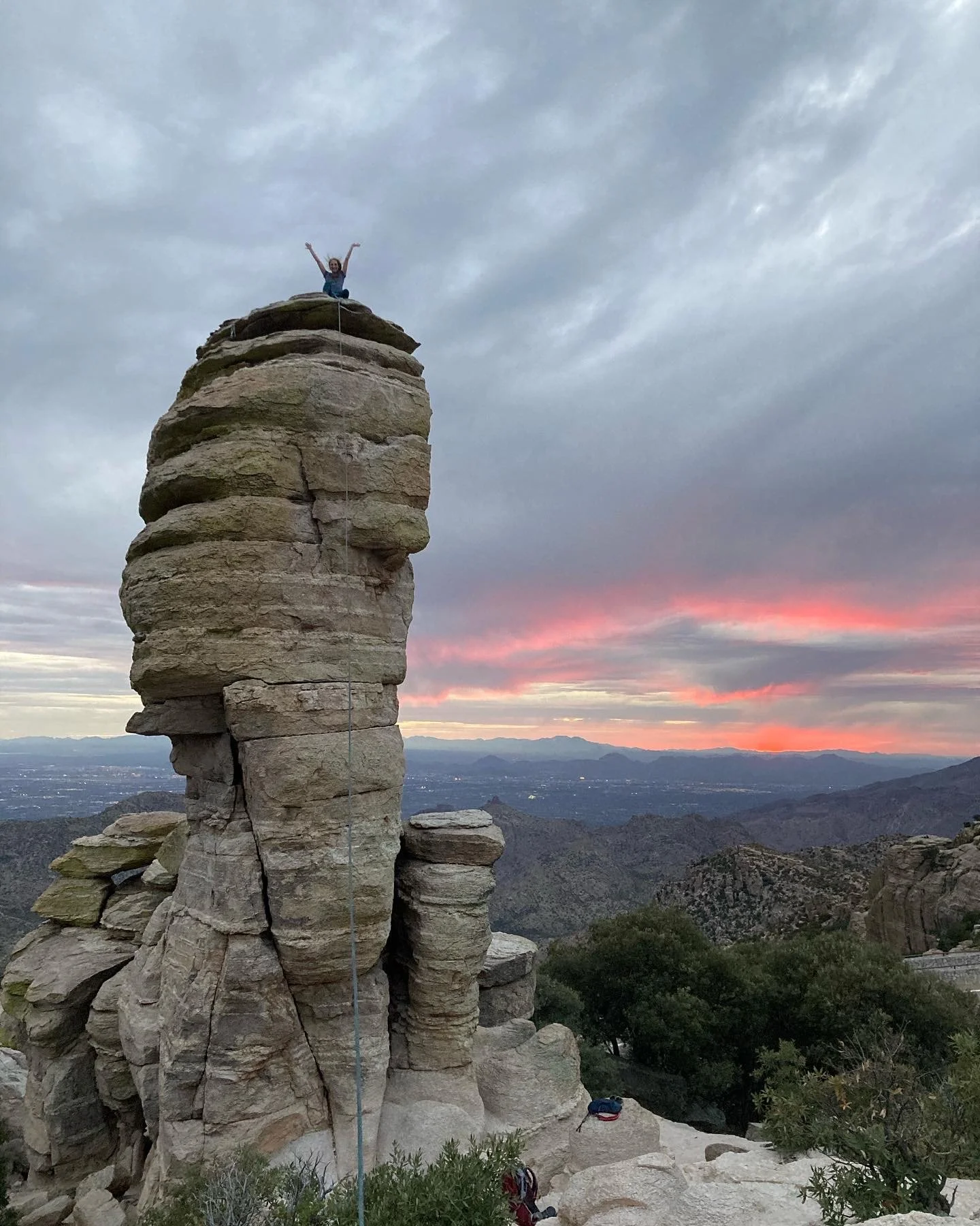 A person standing triumphantly on top of a tall rock formation resembling a human profile with a sunset and mountain range in the background.