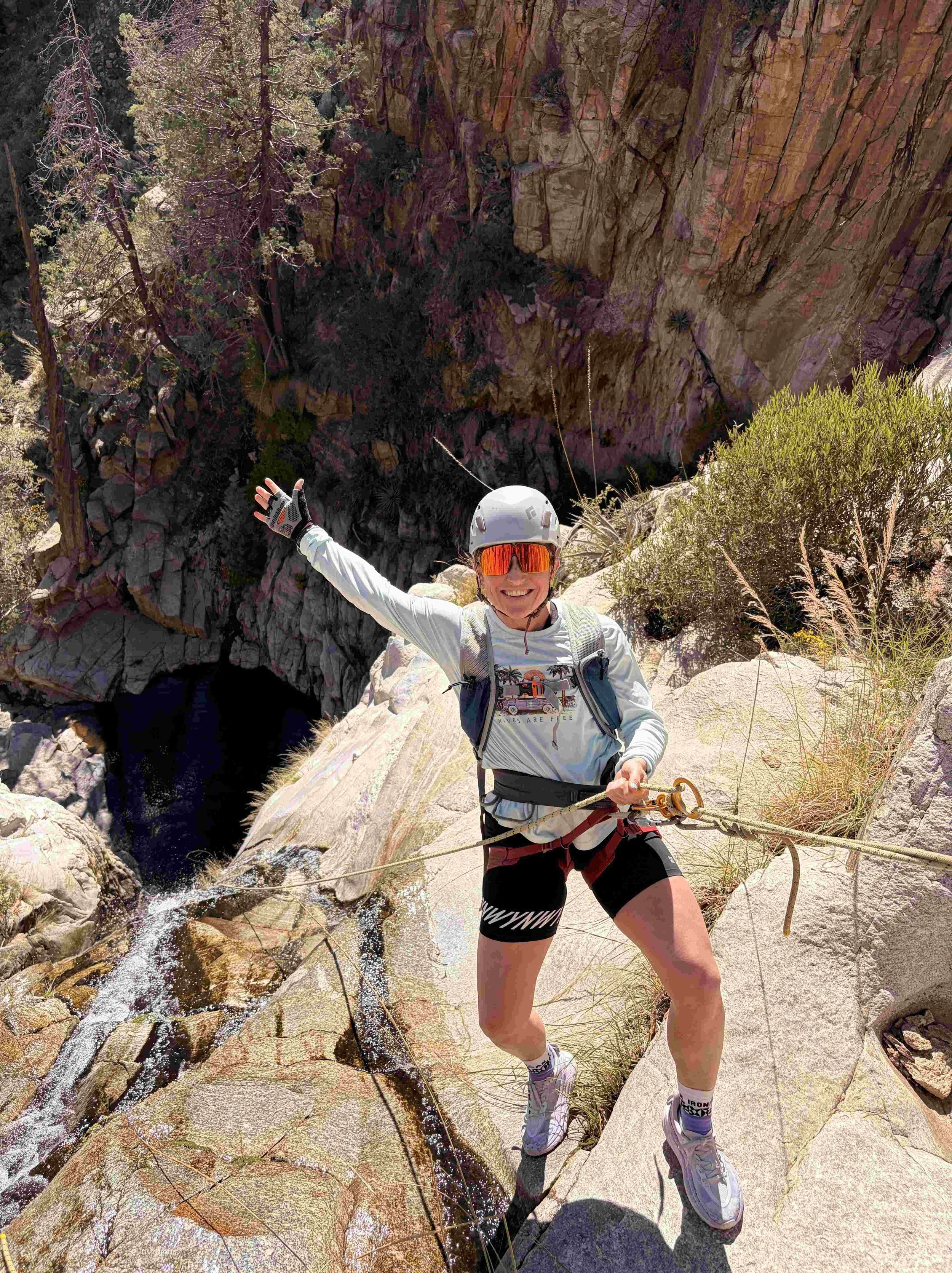 Person wearing a helmet and sunglasses climbing on a rocky cliff with a waterfall in the background.
