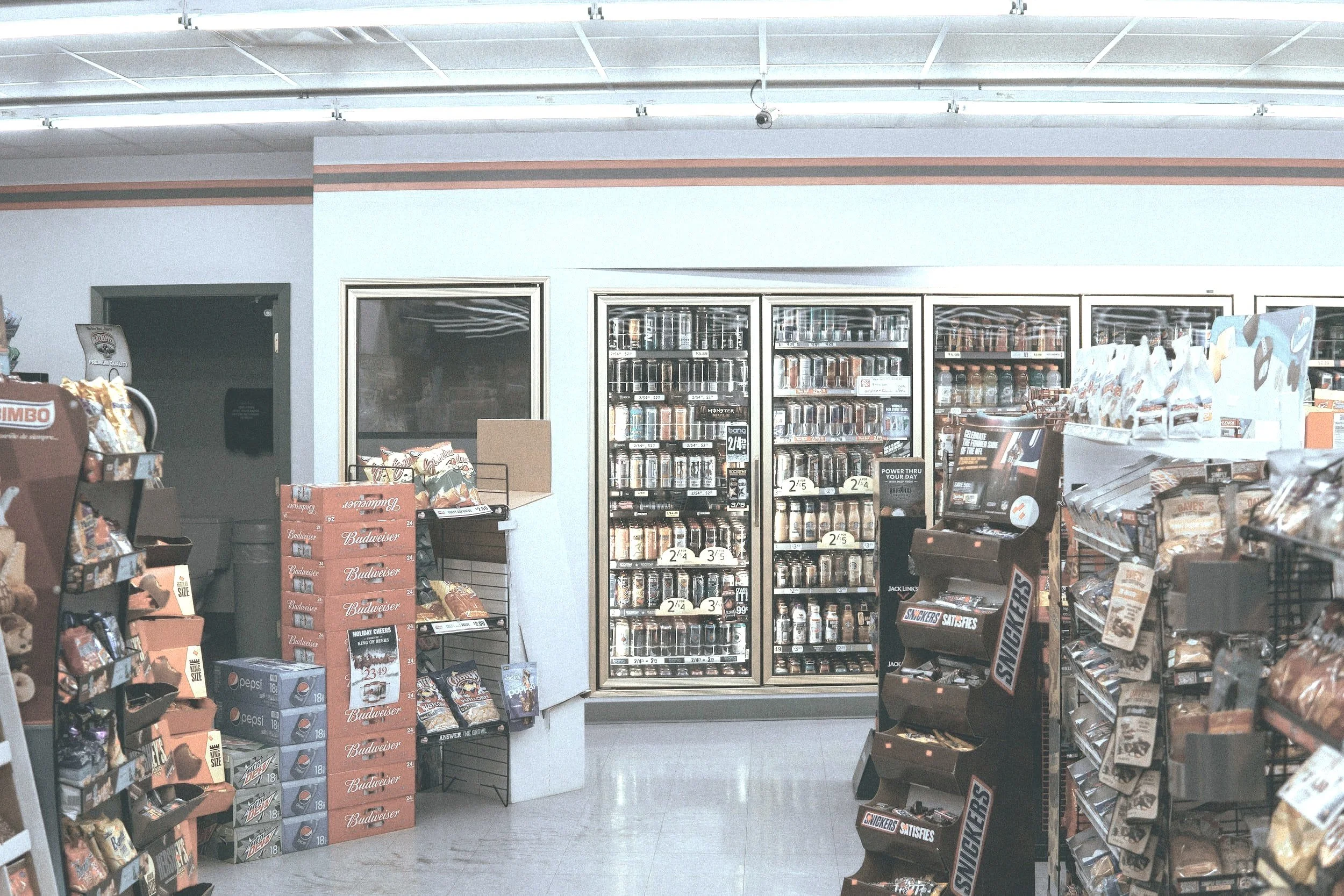 Interior of a convenience store showing shelves with snacks, a refrigerated section with beverages, and display racks for candy.