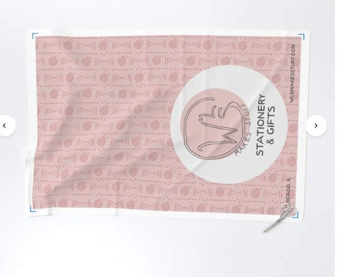 photo of a virtual mockup of a yard of fabric showing a pink pattern of yarn balls, knitting needles, and crochet hooks with the WLS shop logo very large at one end.