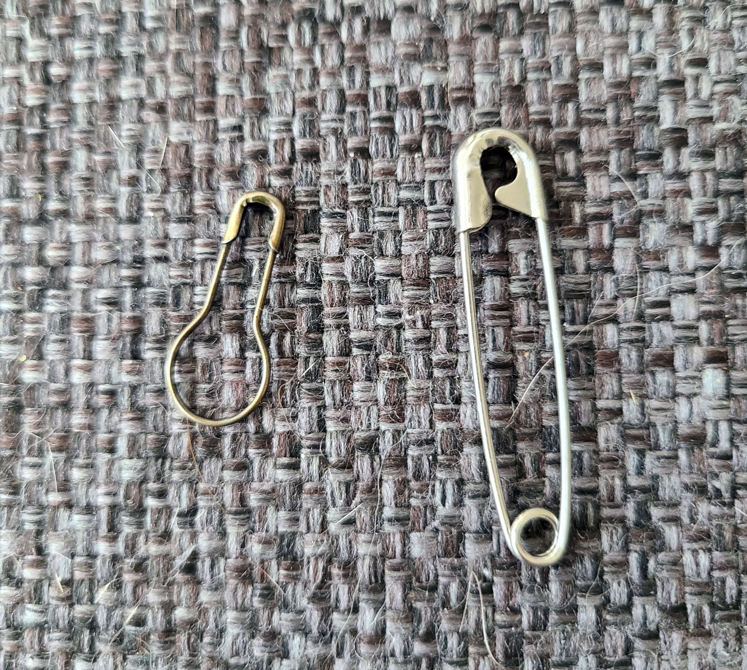 Photo of one coil-less and one coiled safety pin on a gray background. It's clear that the coil-less version is smooth with no areas to catch yarn, while the coiled pin has a coiled spiral at one end and a  lip on the fastener end.