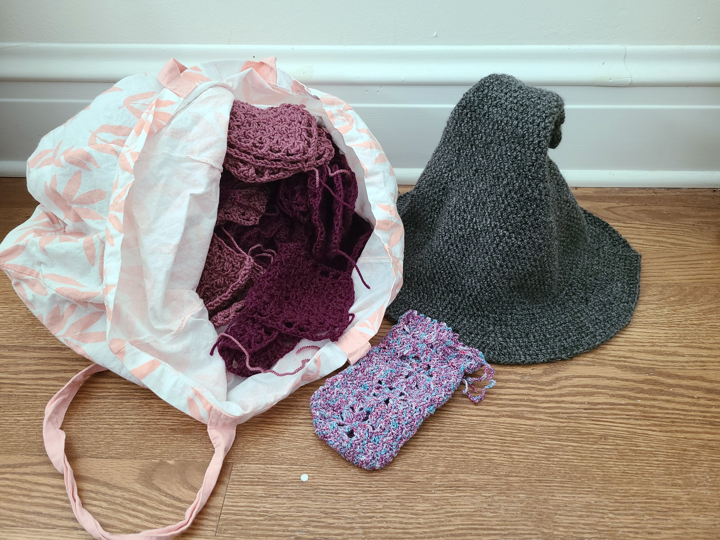 Photo of a bag of crocheted squares, a small crochet pouch, and a crochet witch's hat sitting on a wood floor.