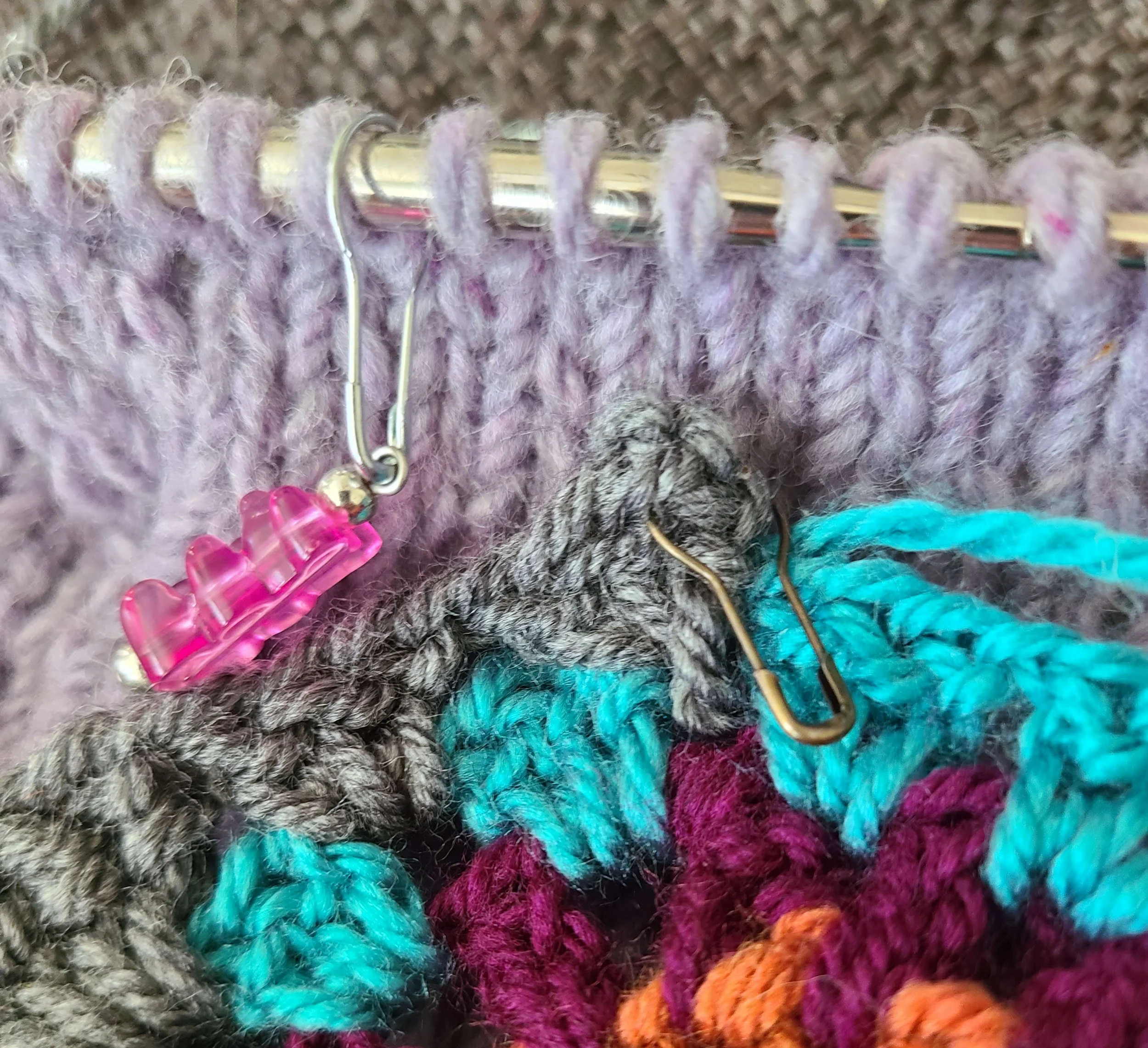 photo of a crochet square held in front of a hand knit project. The crochet square has a coil-less safety pin as a marker; the knit has a stitch marker with a gummy bear shaped bead hanging from a coil-less safety pin closure.
