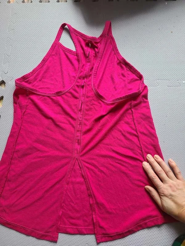 Photo of a bright pink tank top, turned inside out and laid on a light purple background to show the back.. The back is a T-back shape with a split in the bottom middle, and smaller vents in the middle back seam.
