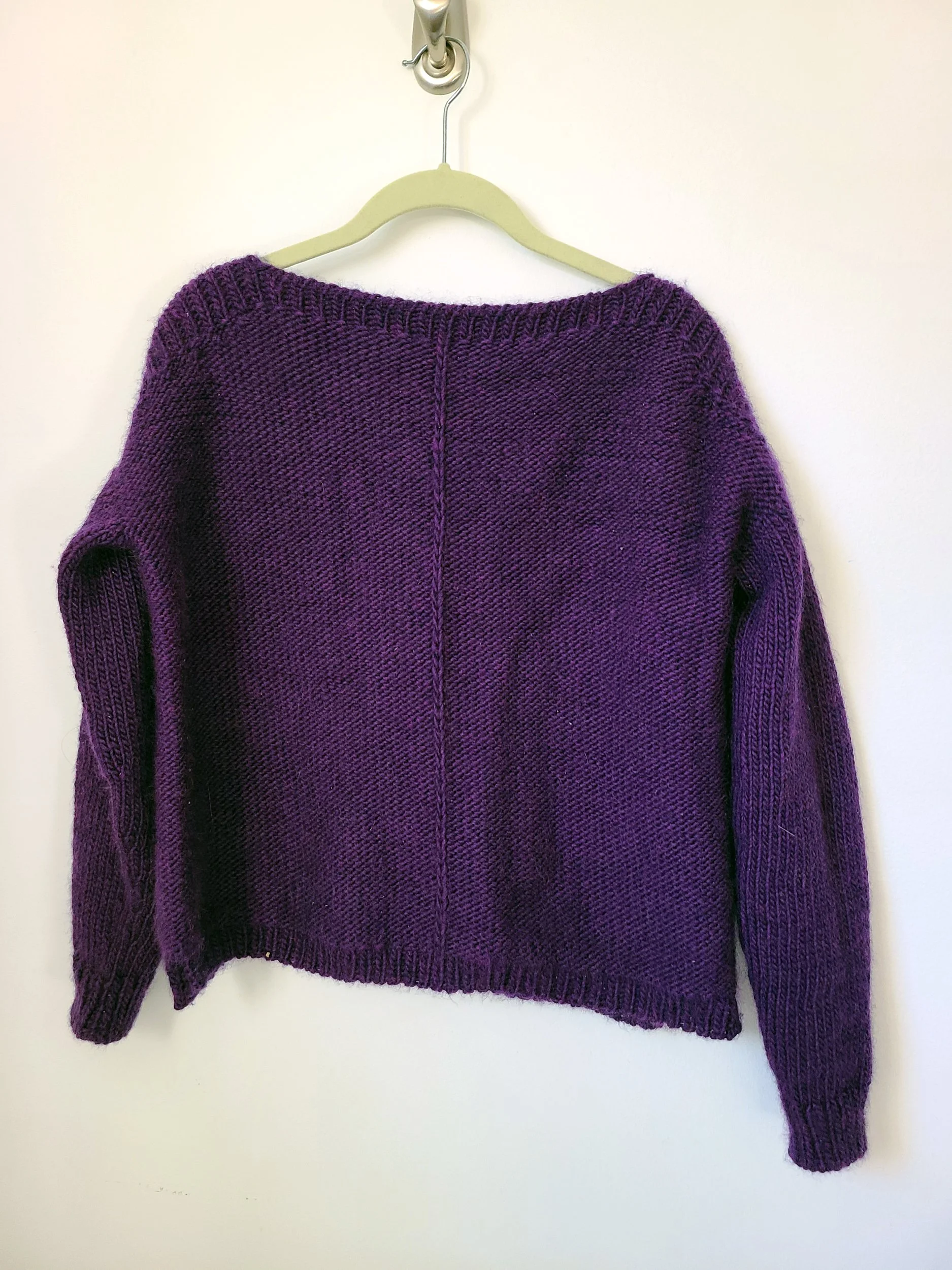Photo of a handknit sweater in dark purple yarn on a hanger in front of a white wall.