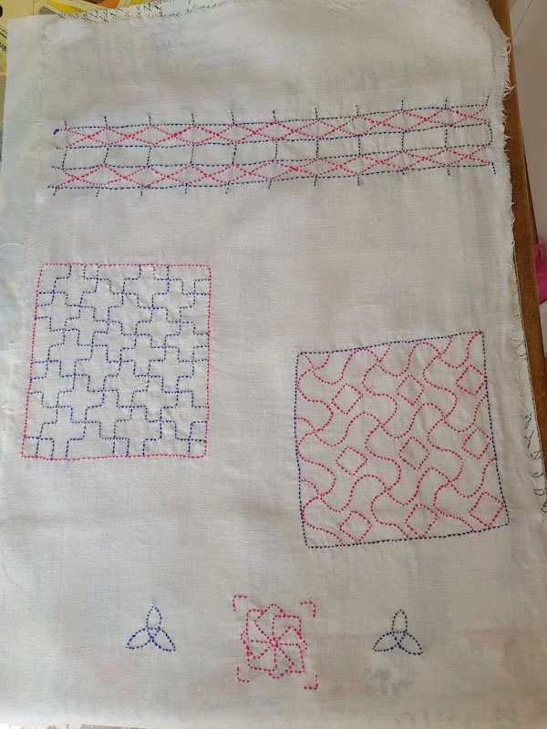Photo of a full rectangular piece of linen on which several different sashiko motifs have been stitched in blue and pink thread.