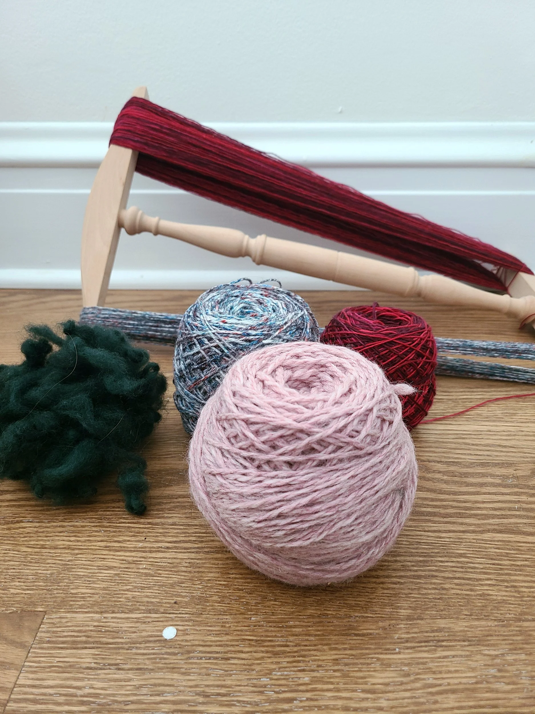 Photo of a ball of pink bulky yarn and a pile of wool locks, a ball of blue speckled sock yarn, and a niddy noddy wrapped in red variegated yarn.