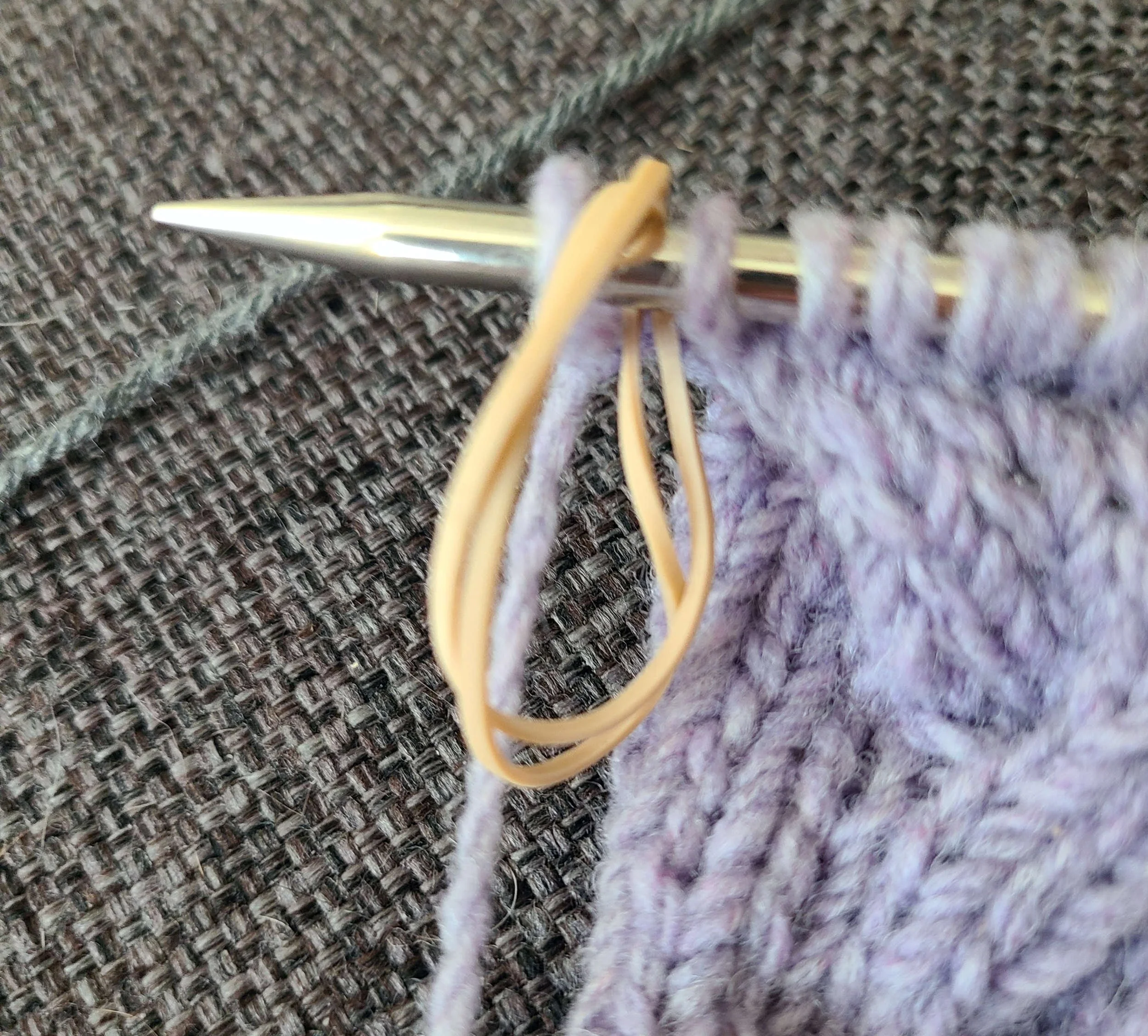 Photo of a hand knit on a needle. A doubled up rubberband is serving as a stitch marker.