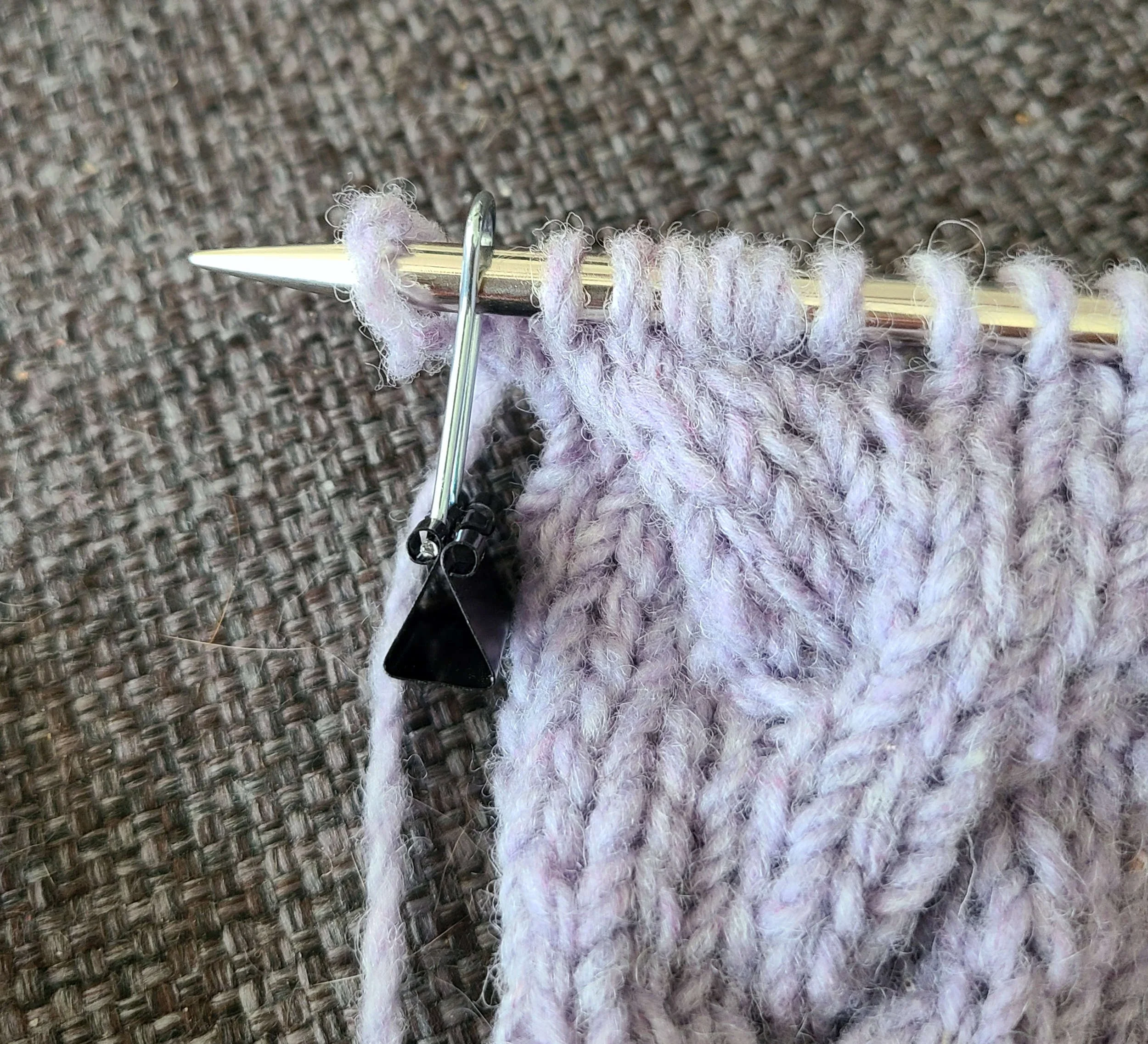 photo of a hand knit project on a knitting needle. A binder clip that is missing one of the metal loops is being used as a ring style stitch marker.