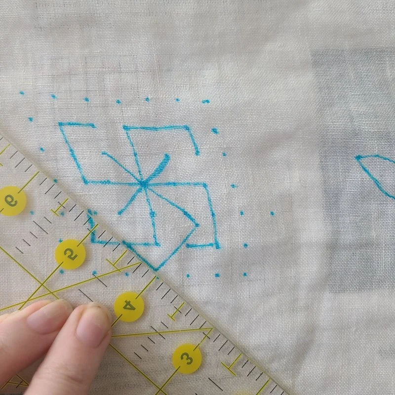 Photo of a piece of linen fabric on which a design has been partially traced in blue marker. A plastic ruler is being used to line up a diagonal line.