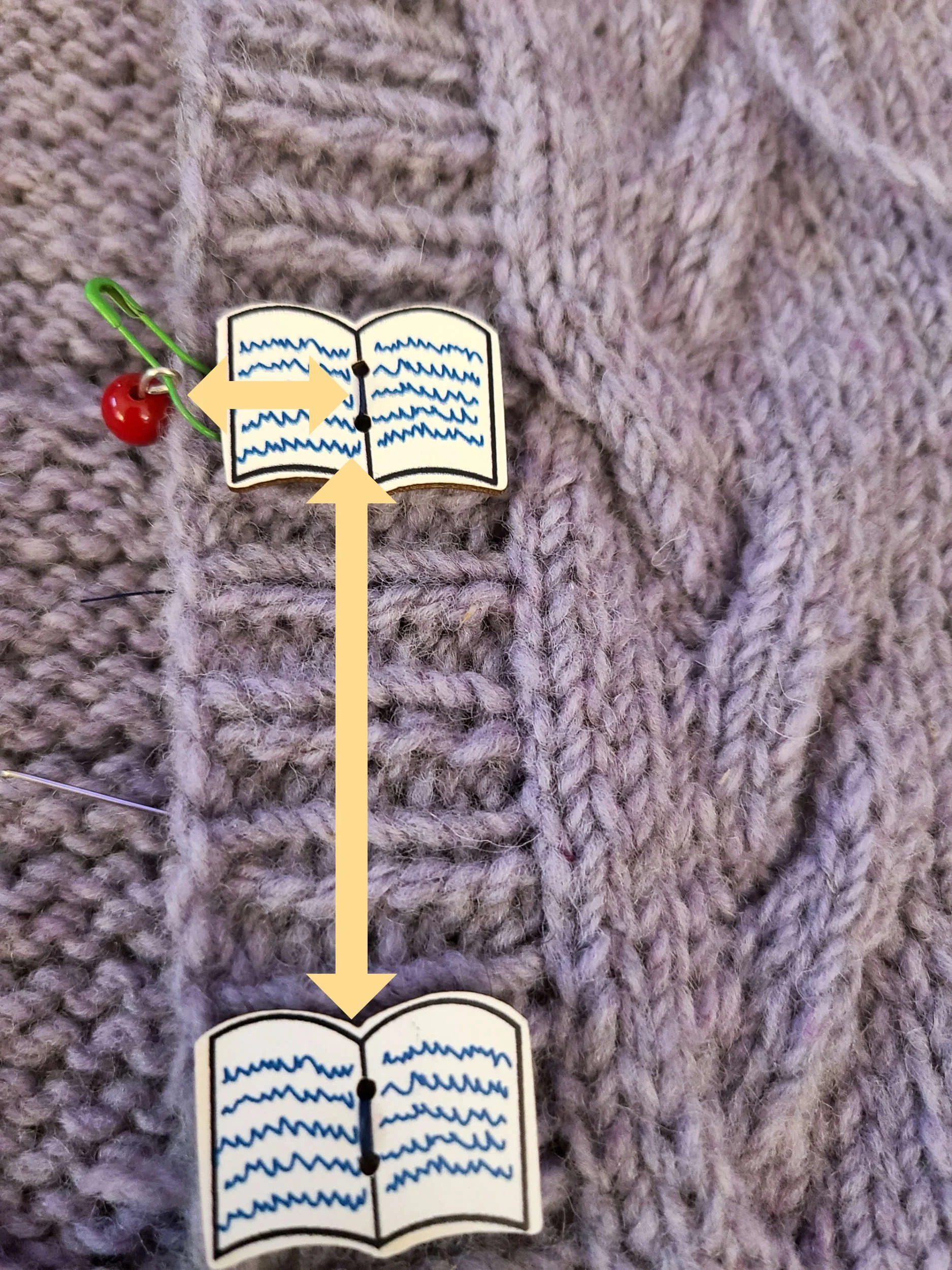 Closeup of a knit cardigan button band. A stitch marker is to the left edge of the button band. The unsewn button has been placed horizontally aligned with the marker but vertically aligned with the sewn button below., shown with arrows.