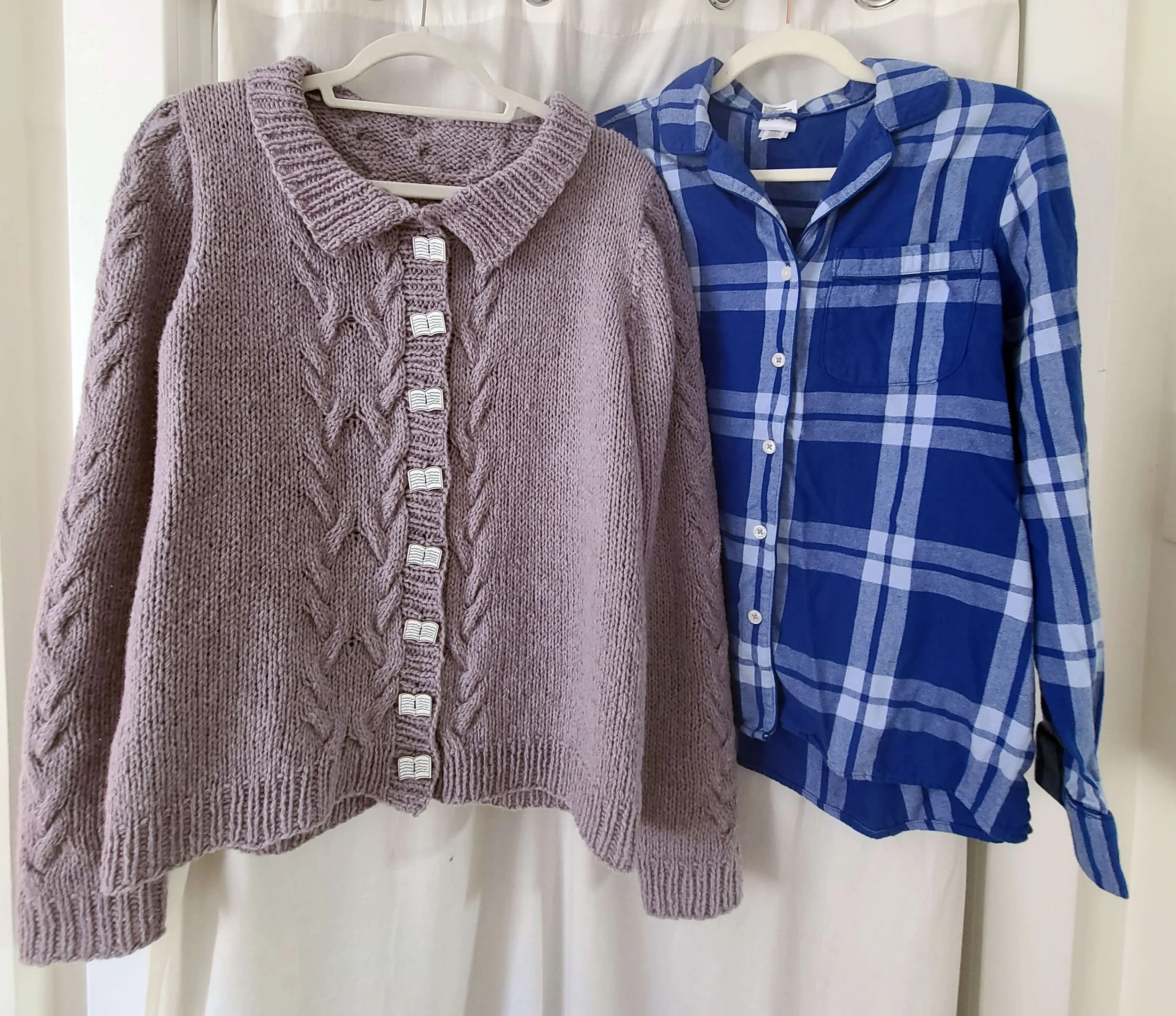 Photo of a knit cardigan and a flannel shirt with buttons attached and aligned, on hangers in front of a white curtain.