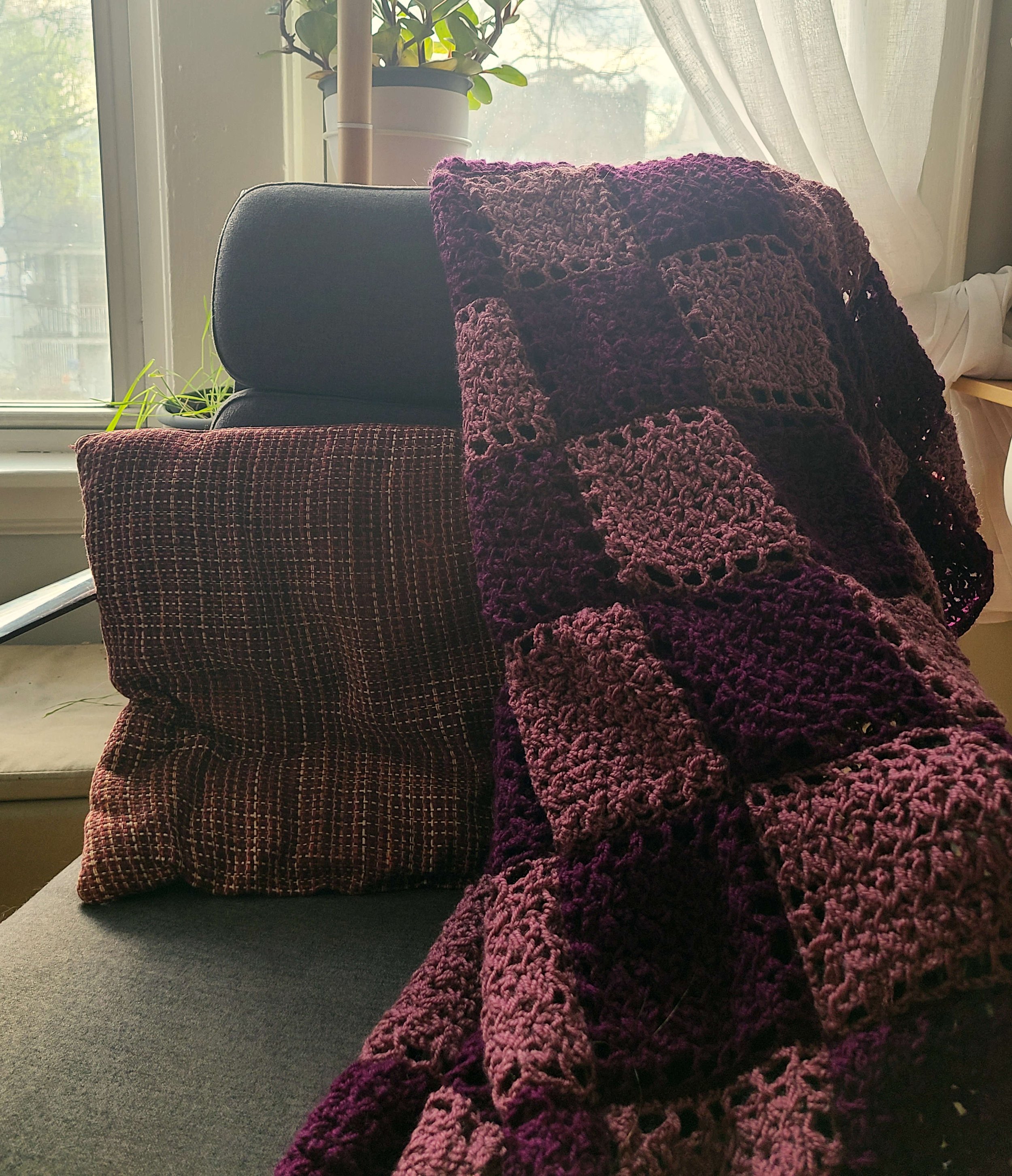 photo of an afghan consisting of light and dark purple crocheted squares draped over a desk chair with a matching throw pillow, in front of a window.