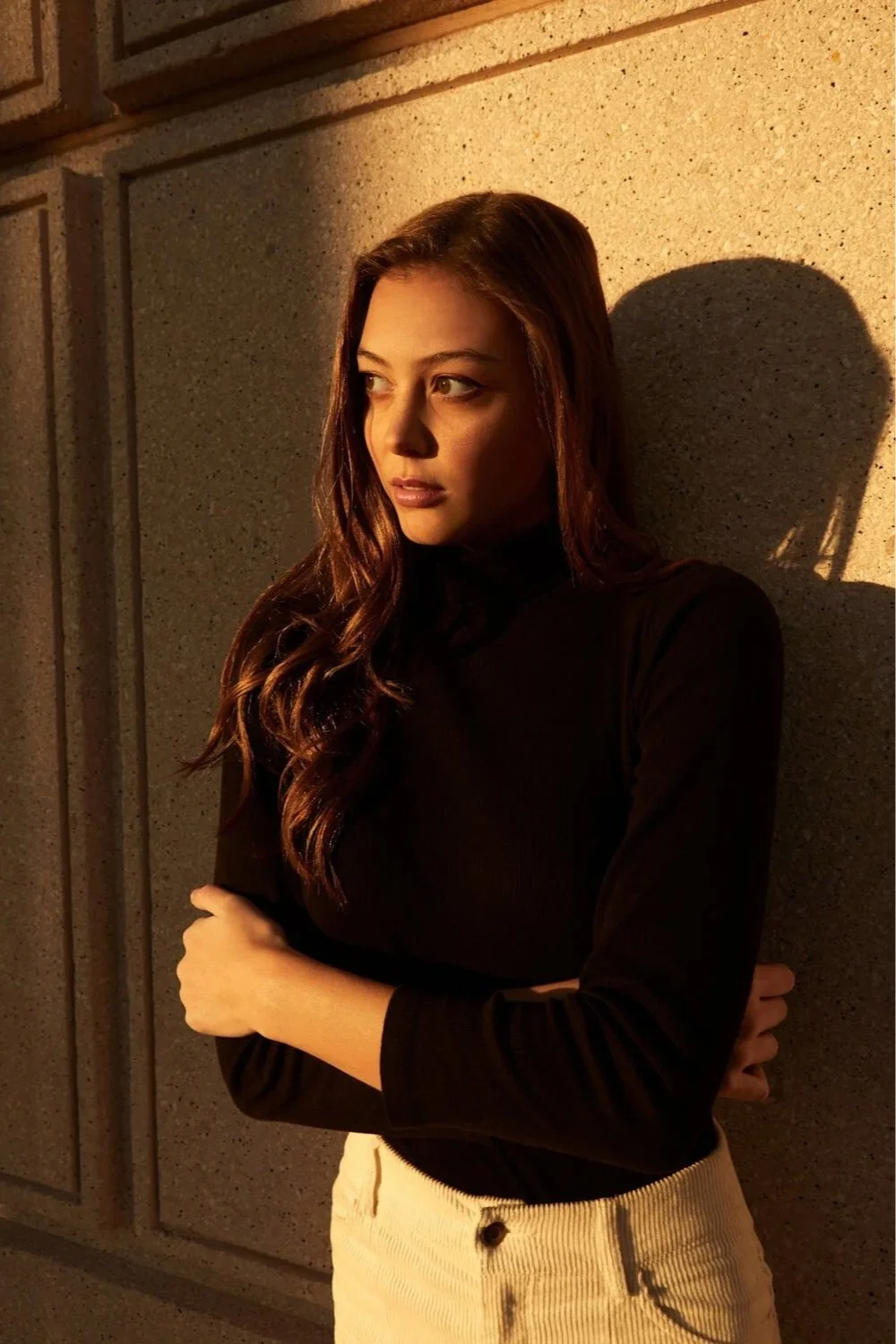 Facetune App Ad - A young woman with wavy brown hair wearing a black turtleneck and beige pants, standing against a concrete wall with warm lighting casting shadows.