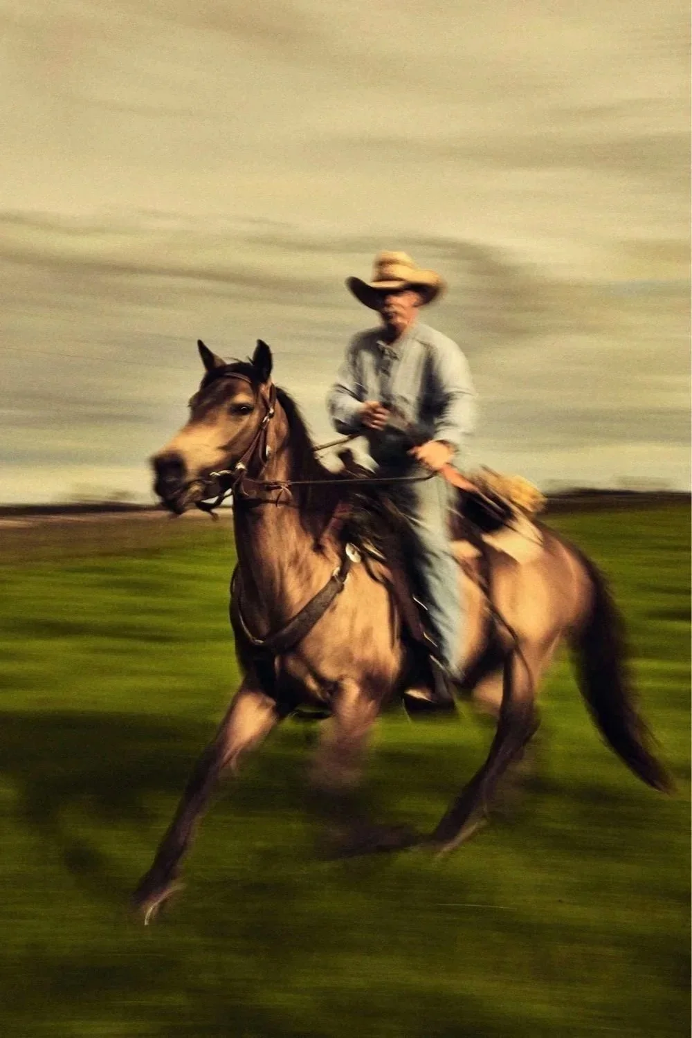 A person wearing a cowboy hat riding a running horse across a grassy field during a cloudy day.